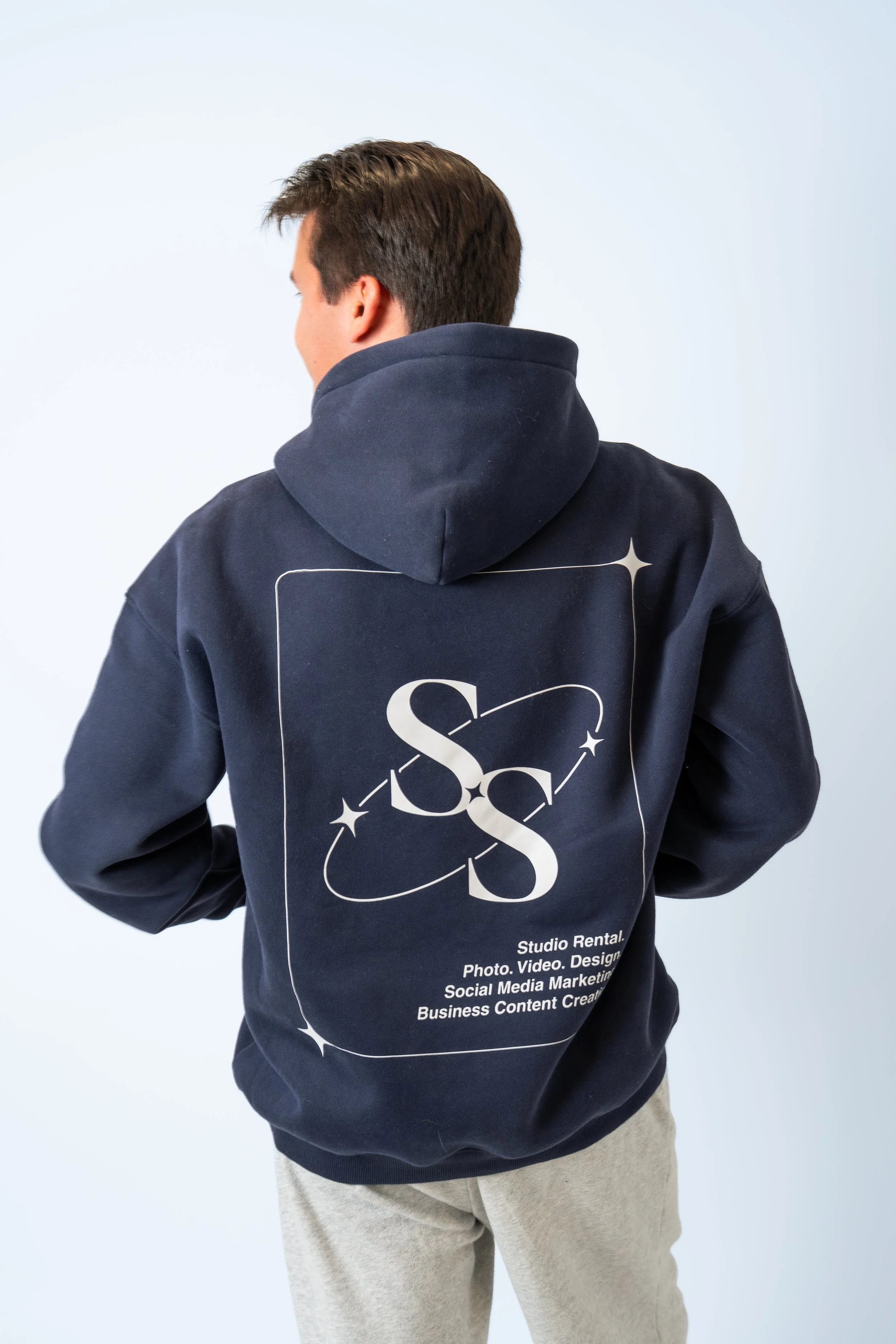 Back of a man wearing a navy hoodie with white graphic and text, against a light background.