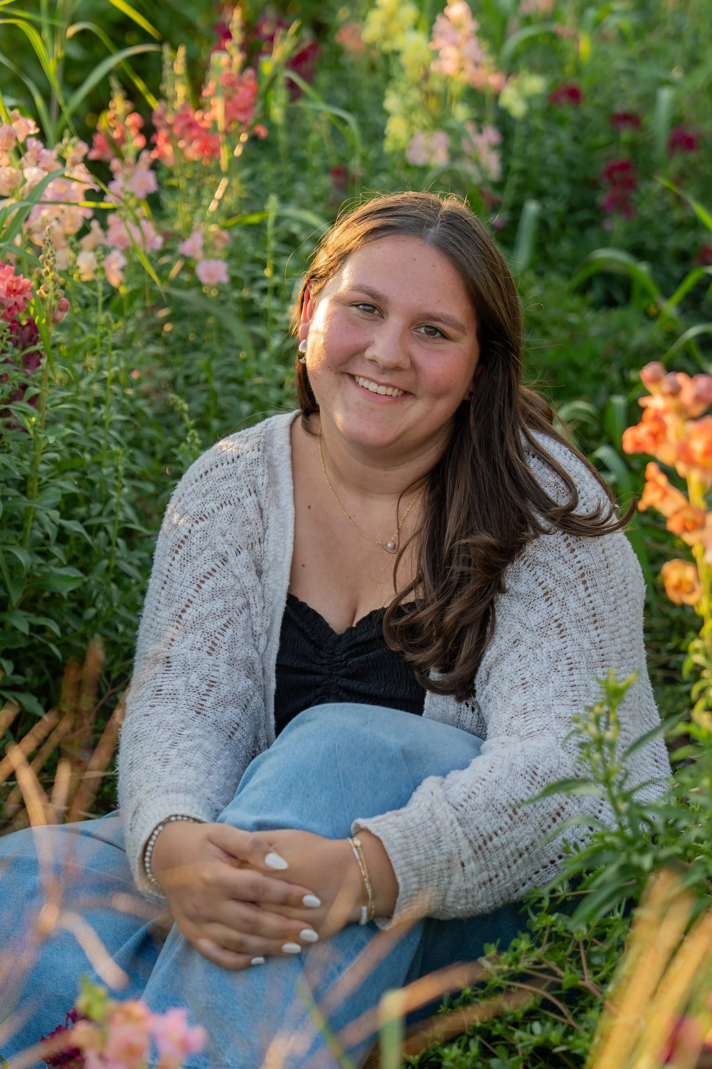 A young woman sitting amidst colorful flowers and green foliage, smiling at the camera.