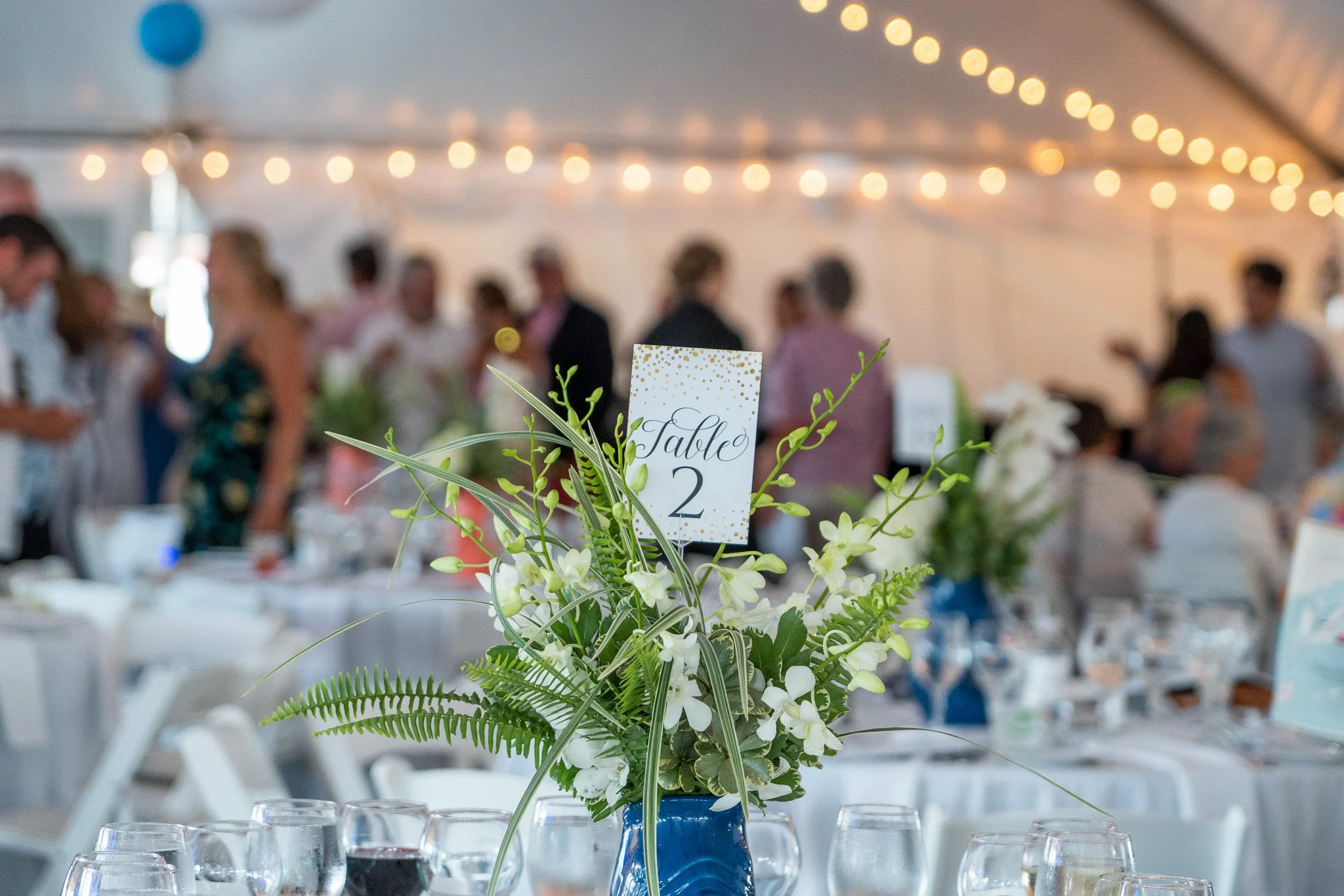 Table centerpiece with white flowers and green foliage, labeled 'Table 2' in gold and black, set for a wedding or celebration with blurred guests and string lights in background.