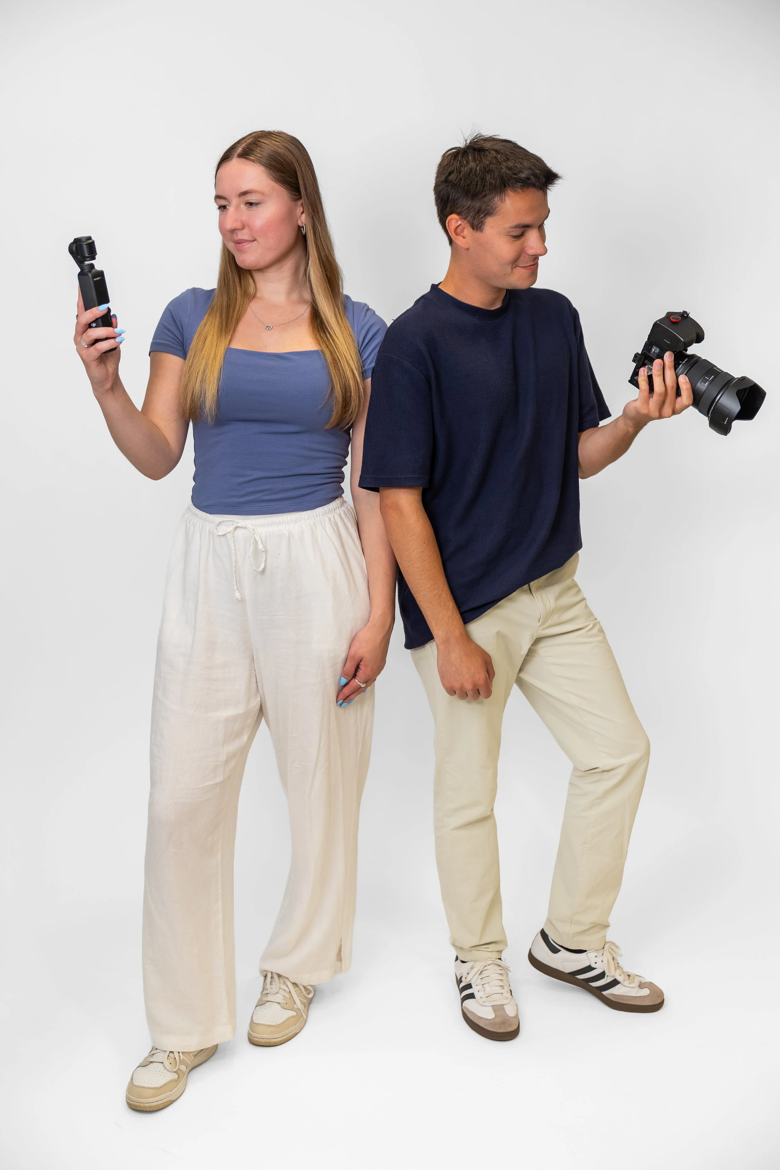 Two young adults standing back-to-back against a white background, each holding a camera and looking down at their respective devices.