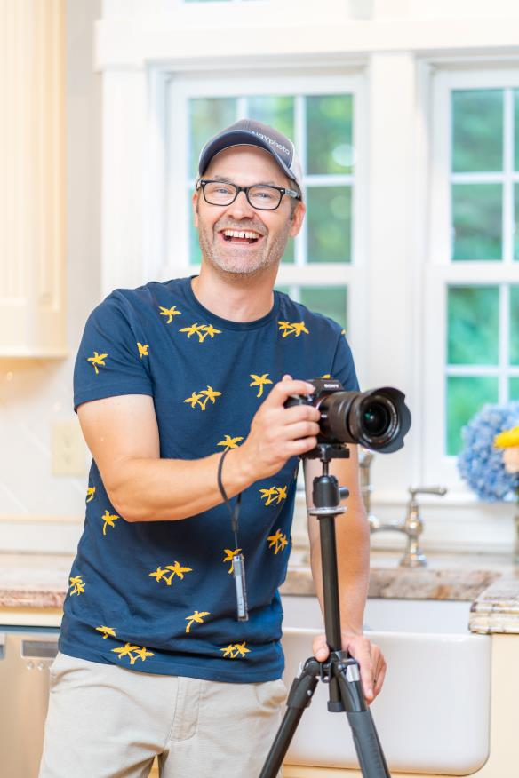 A smiling man in a blue shirt with yellow bird patterns wearing glasses and a cap, holding a camera on a tripod inside a bright kitchen with large windows and a vase of flowers.