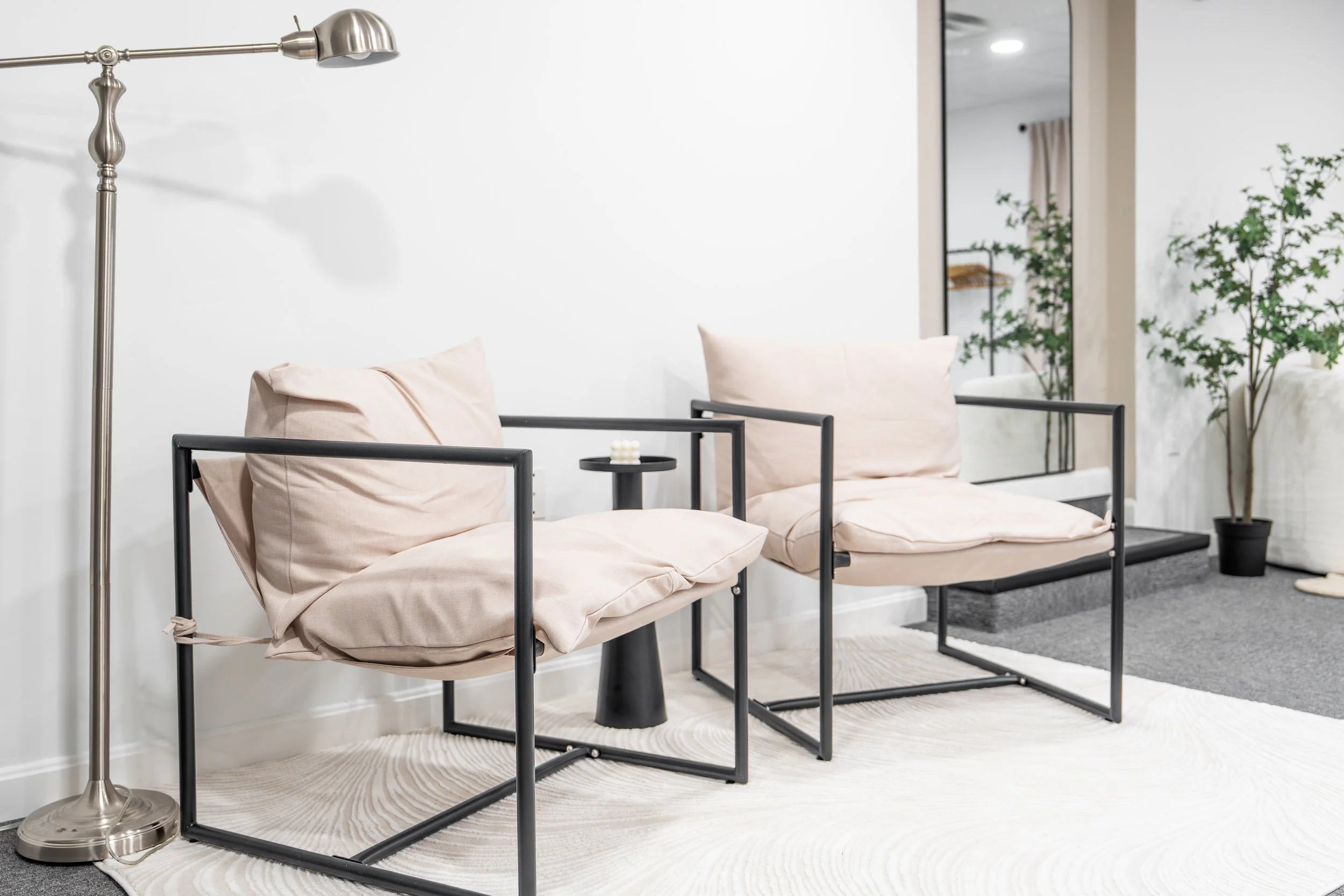 Modern waiting area with two beige cushioned chairs with black metal frames, a small black side table with a white object on it, a tall silver floor lamp, a large mirror, and potted green plants in the background.