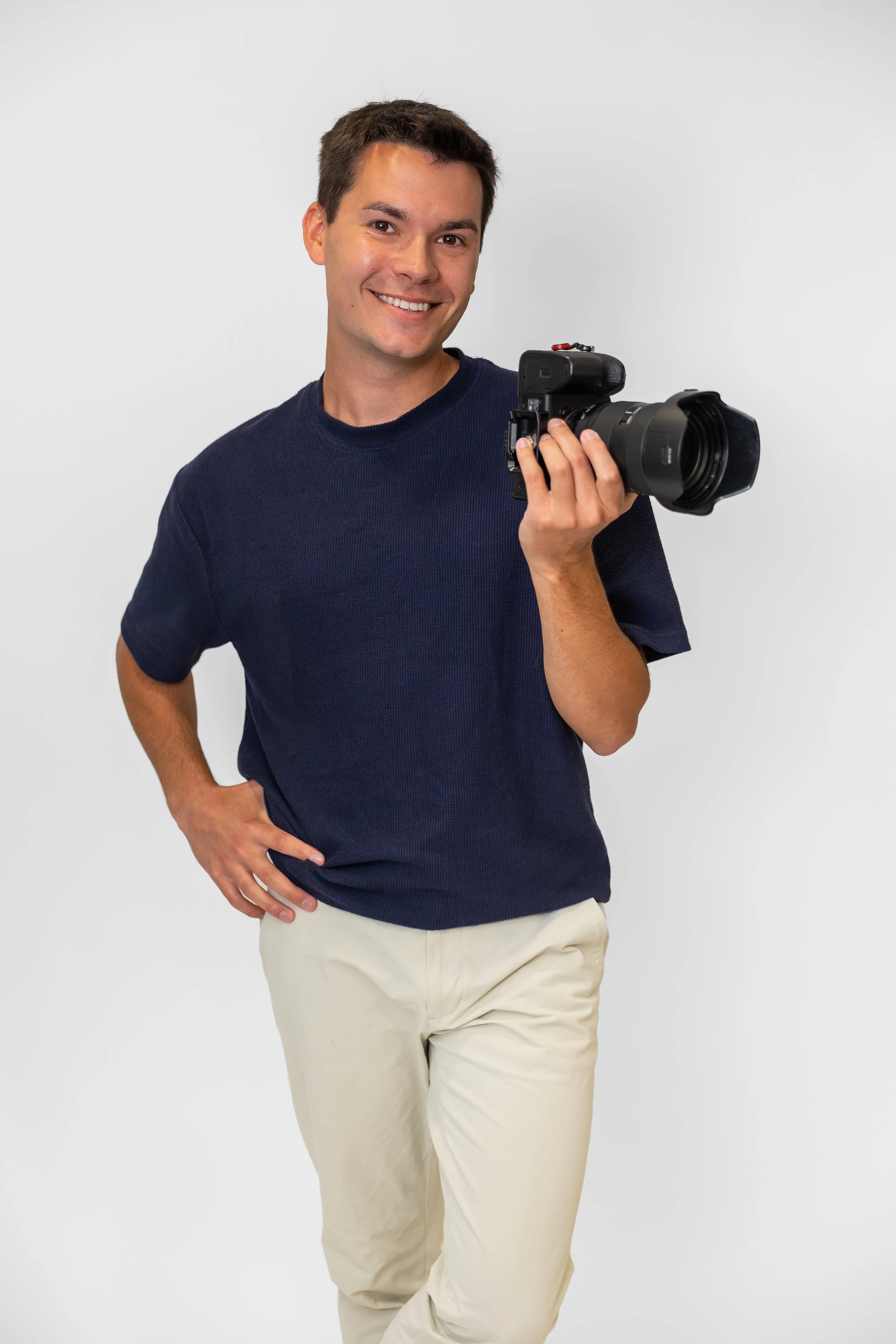 Young man in a navy blue t-shirt and beige pants holding a camera, smiling and posing against a plain white background.