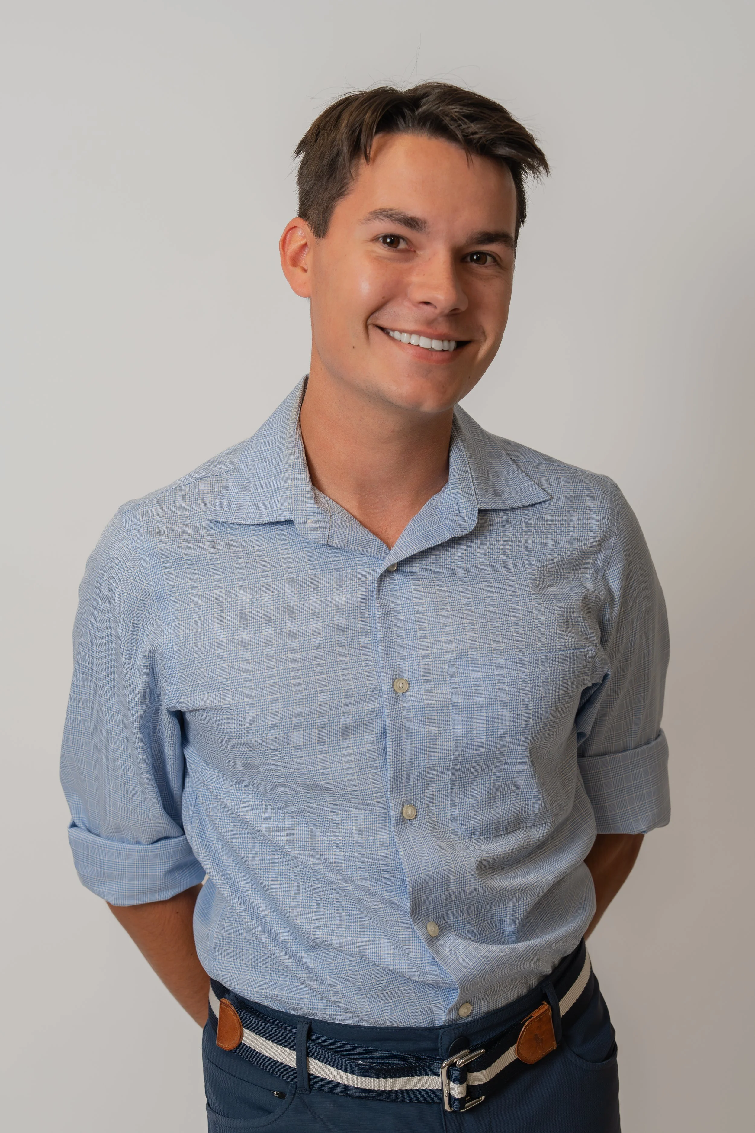 Portrait of a young man with short brown hair, smiling, wearing a light blue checkered shirt with rolled-up sleeves, and navy blue pants with a striped belt, standing against a plain white background.