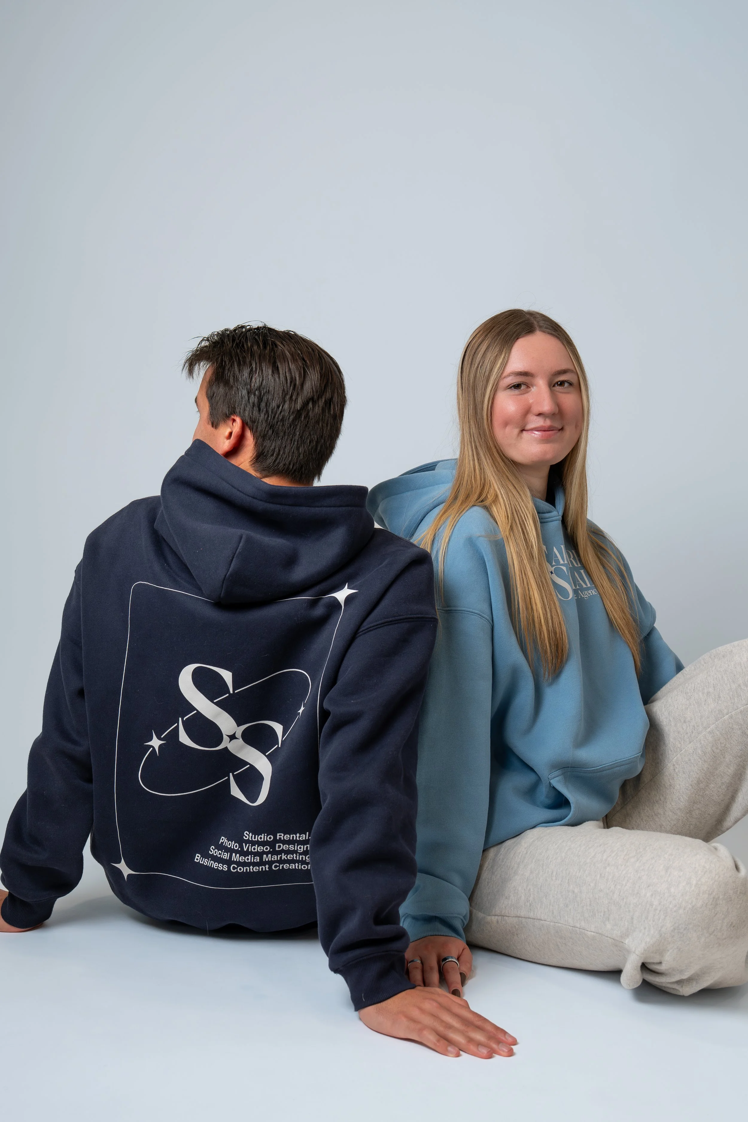 A young man and woman are sitting on the floor against a plain light background. The man, facing away, wears a navy hoodie with a logo on the back. The woman, facing forward with a slight smile, wears a light blue hoodie and beige sweatpants.