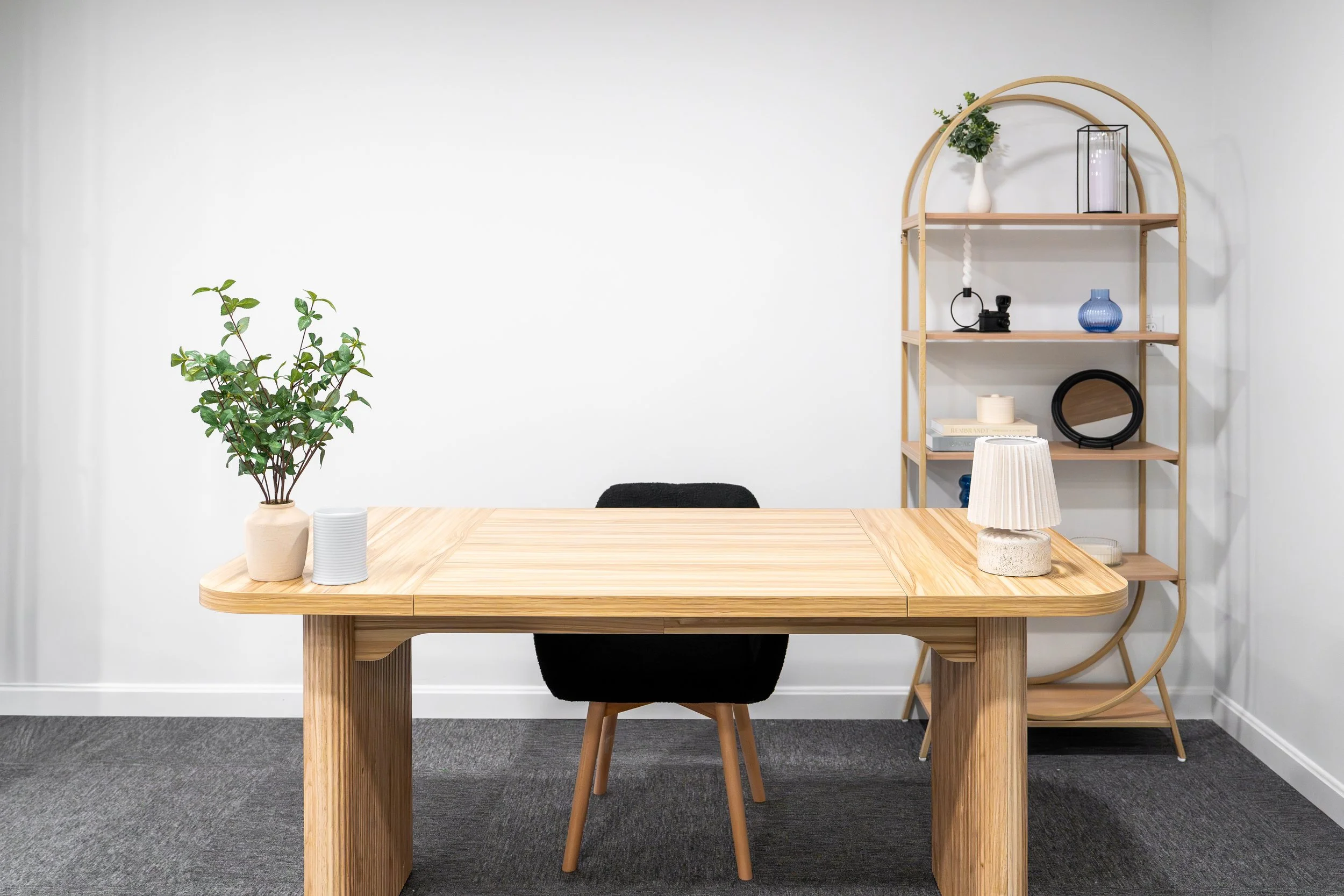 Minimalist office with a wooden table, black chair, two white vases with green plants, a lamp, and a wooden shelf with decorative items against a plain white wall.