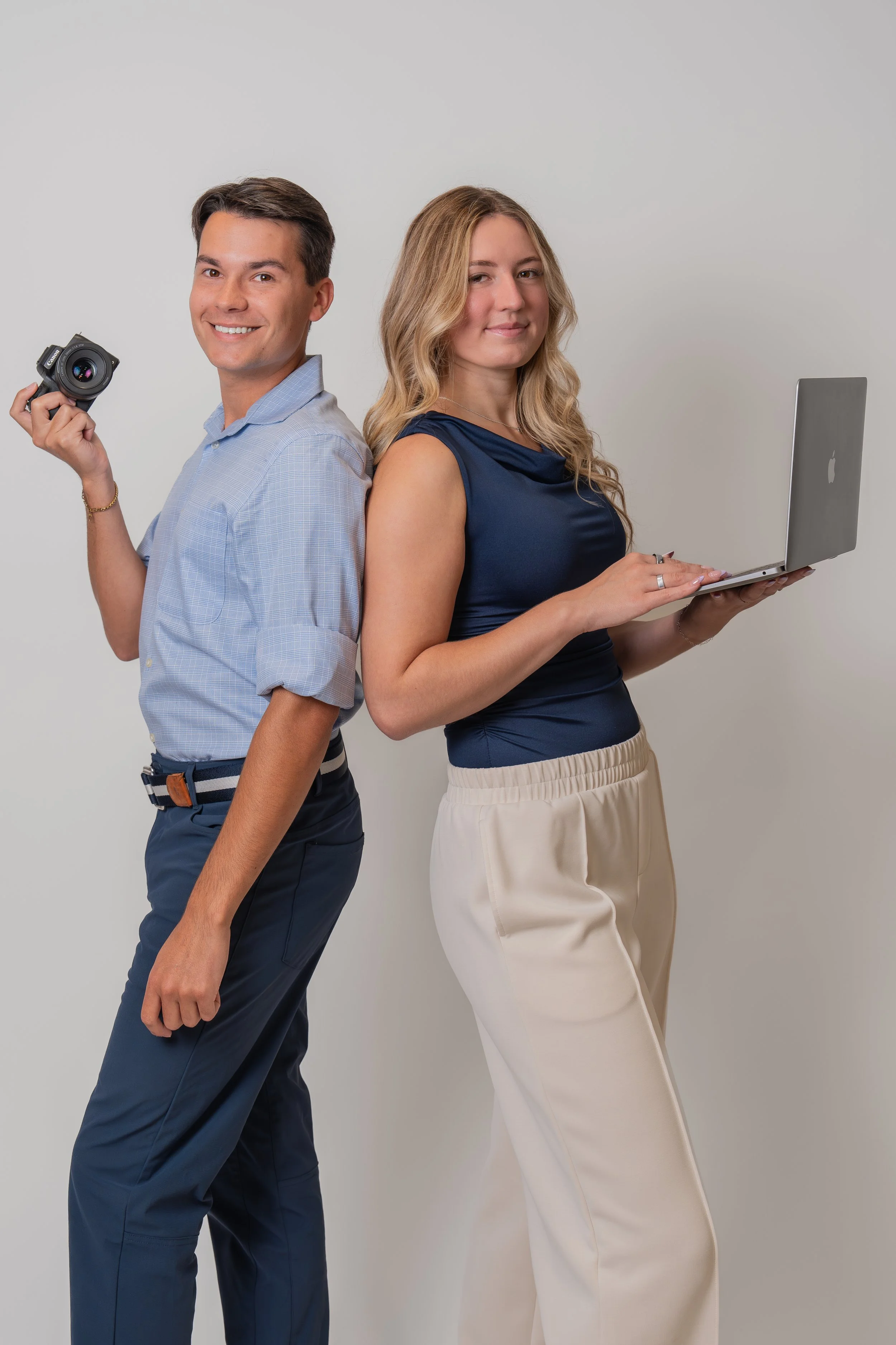 A man and a woman standing back to back, smiling at the camera. The man holds a camera in his right hand, and the woman is using a laptop. They are dressed in business casual clothing against a plain white background.