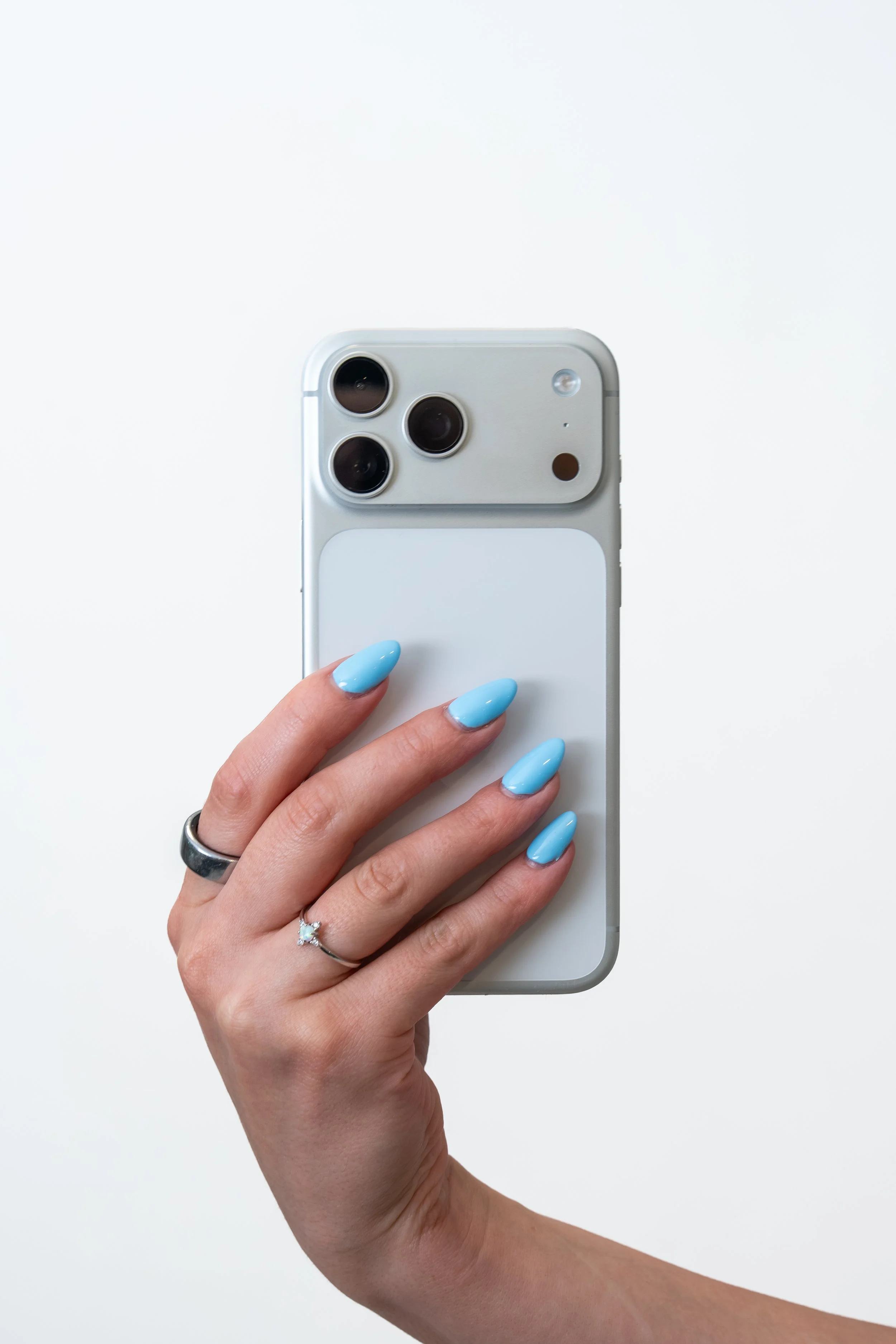 Close-up of a hand with light blue manicured nails holding a silver iPhone with a triple camera system and flash, against a plain white background.