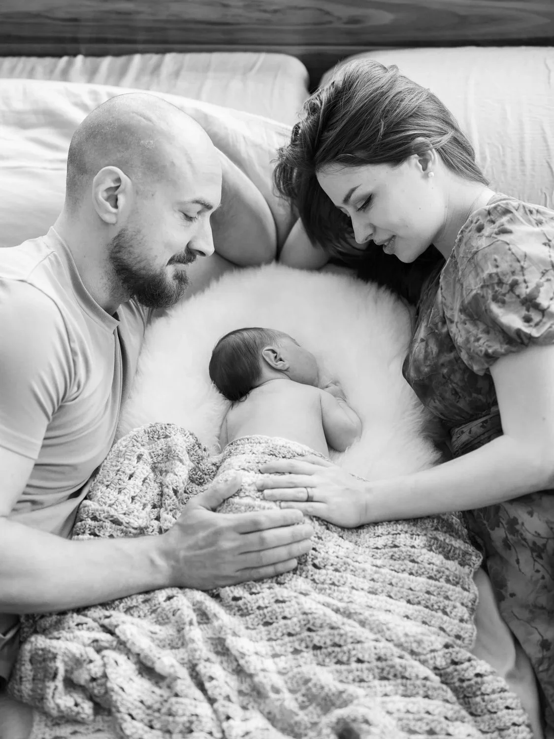 Newborn lifestyle black and white photo of a man and a woman laying on a bed with a newborn between them. 