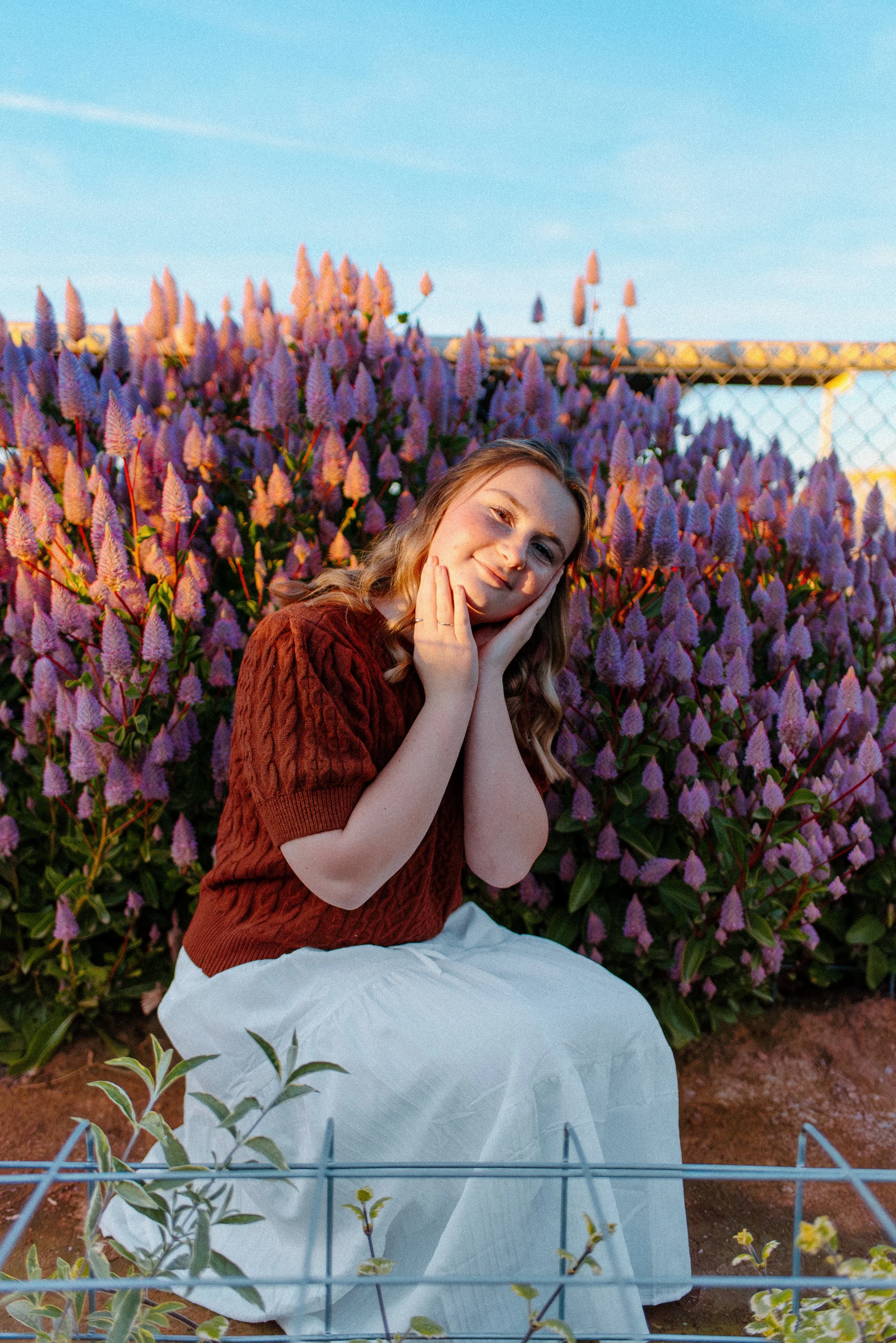 A young woman with wavy blonde hair wearing a brown sweater and white skirt, sitting in front of a large bush of pink and purple flowers, smiling gently with hands on her face, during sunset.