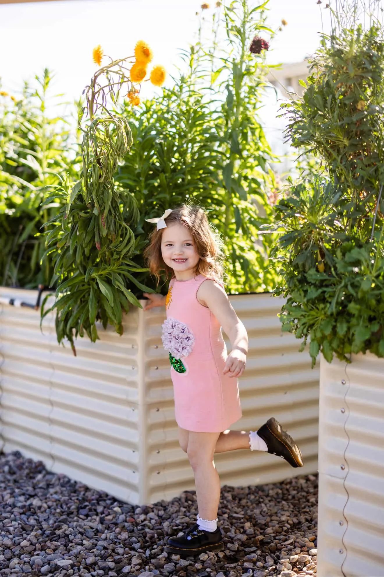 Young girl with brown hair in a pink dress smiling and playing outdoors among green plants and tall flowers in a raised garden bed.
