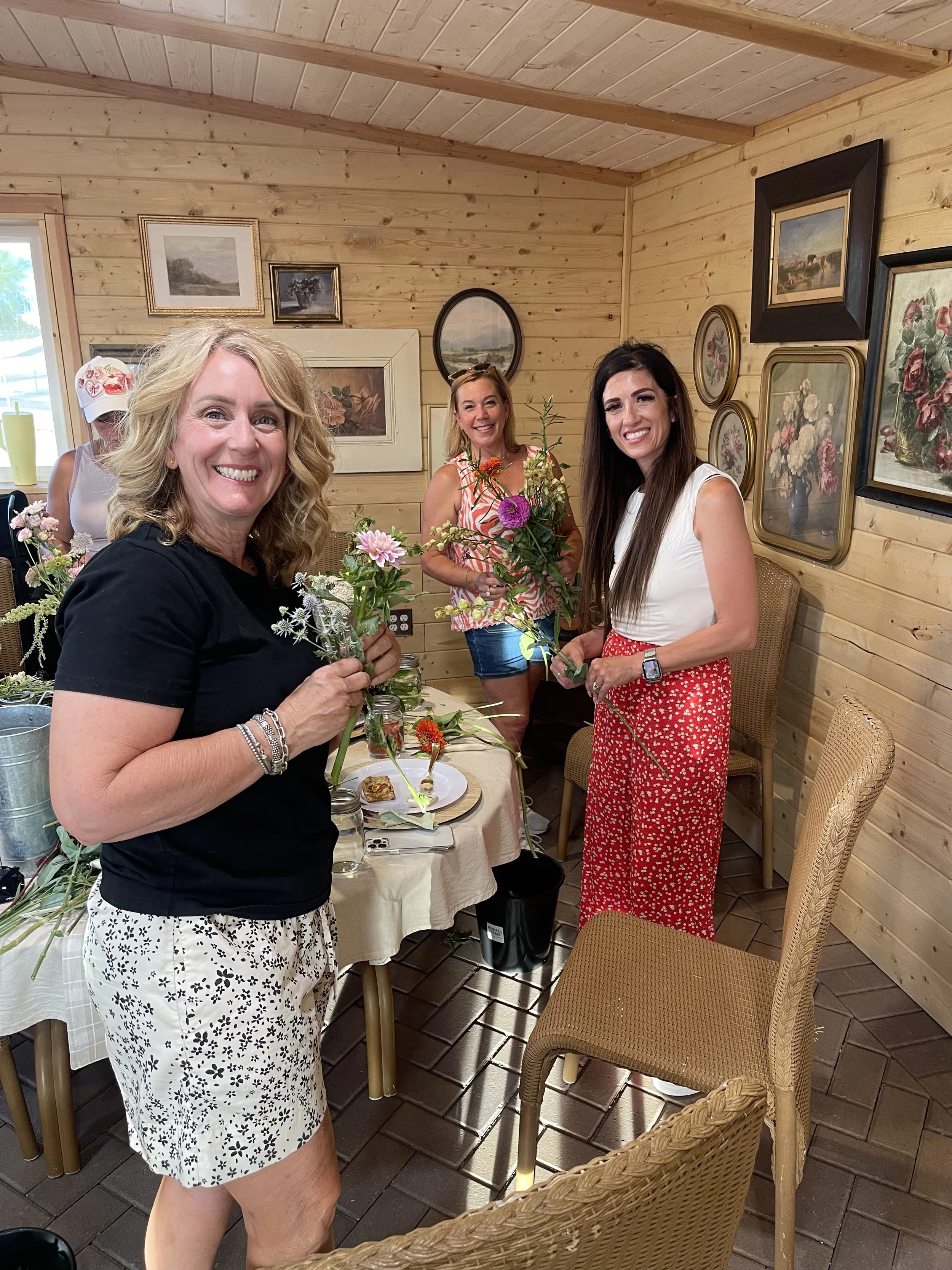 Three women standing around a table with flowers, smiling, in a wooden room with picture frames on the wall.