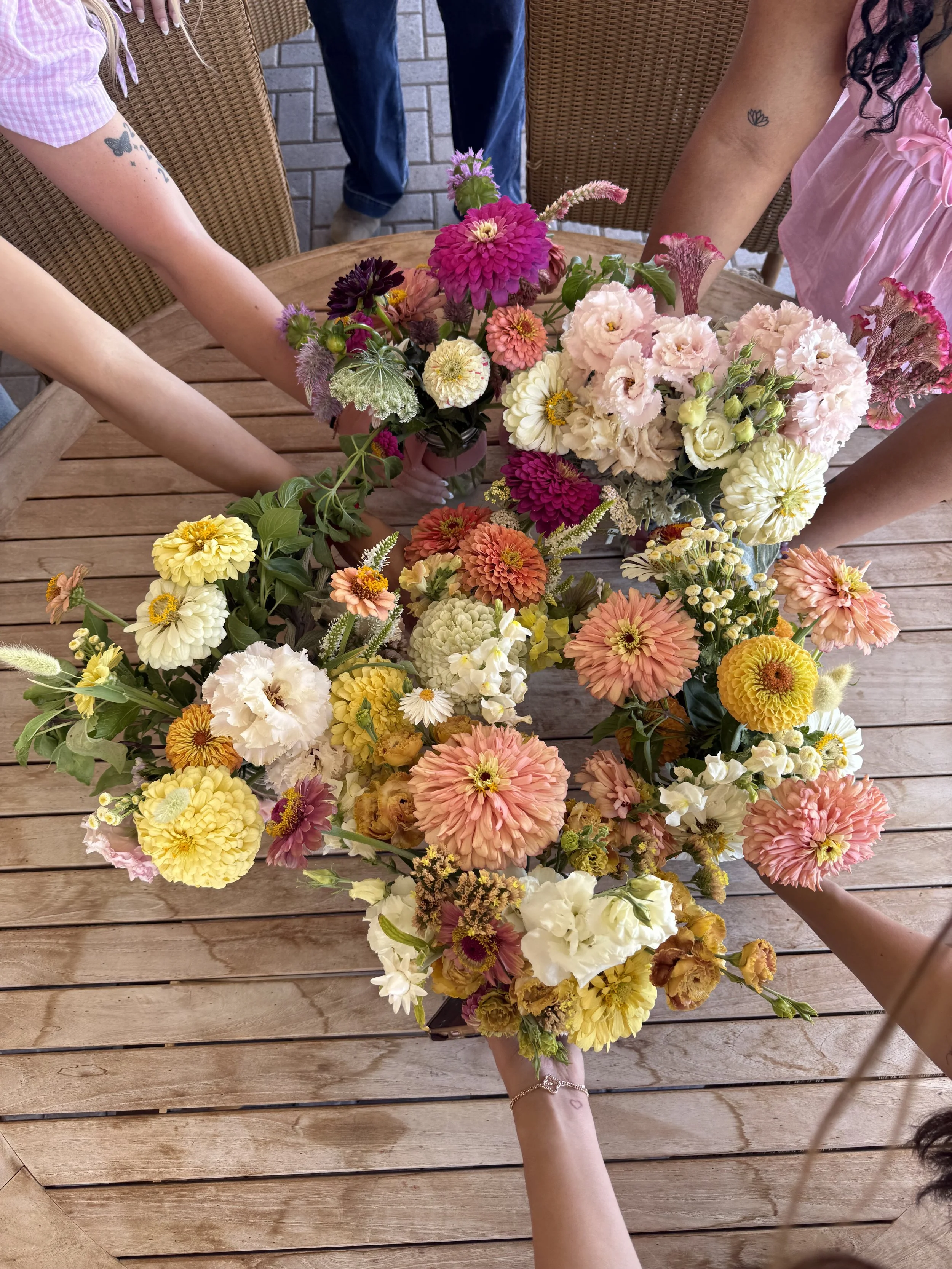 Many colorful flowers arranged on a round wooden table, with four people reaching towards the flowers.