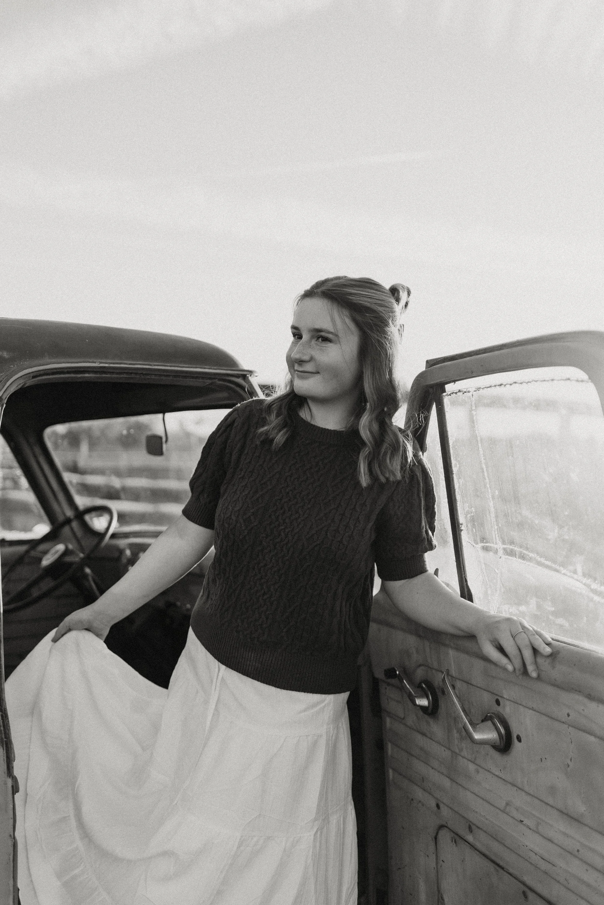 A young woman standing next to a vintage car, holding her skirt, and smiling outdoors.