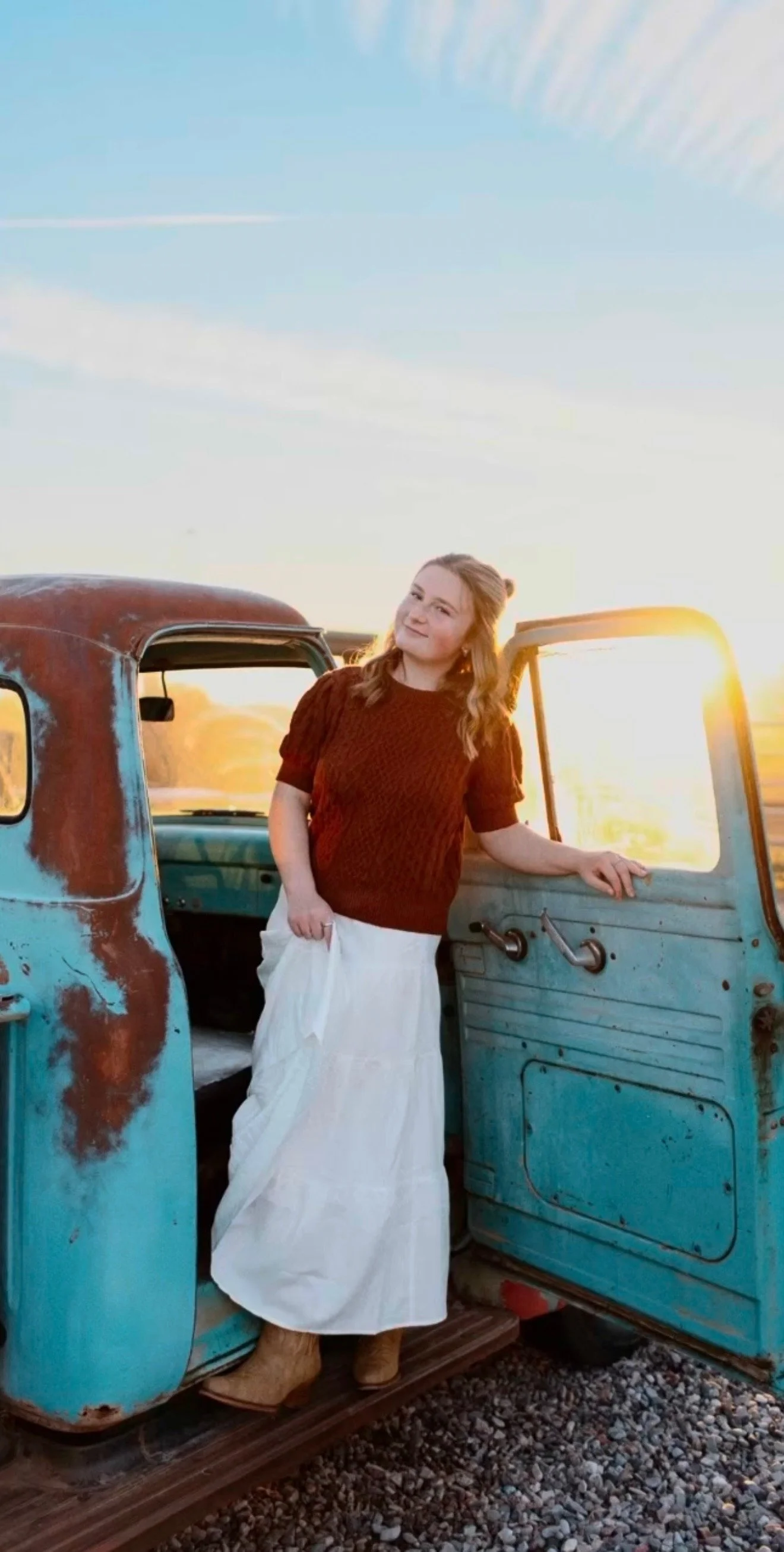 A woman standing outside a vintage blue truck at sunset, wearing a red sweater, a white skirt, and tan shoes, smiling slightly.
