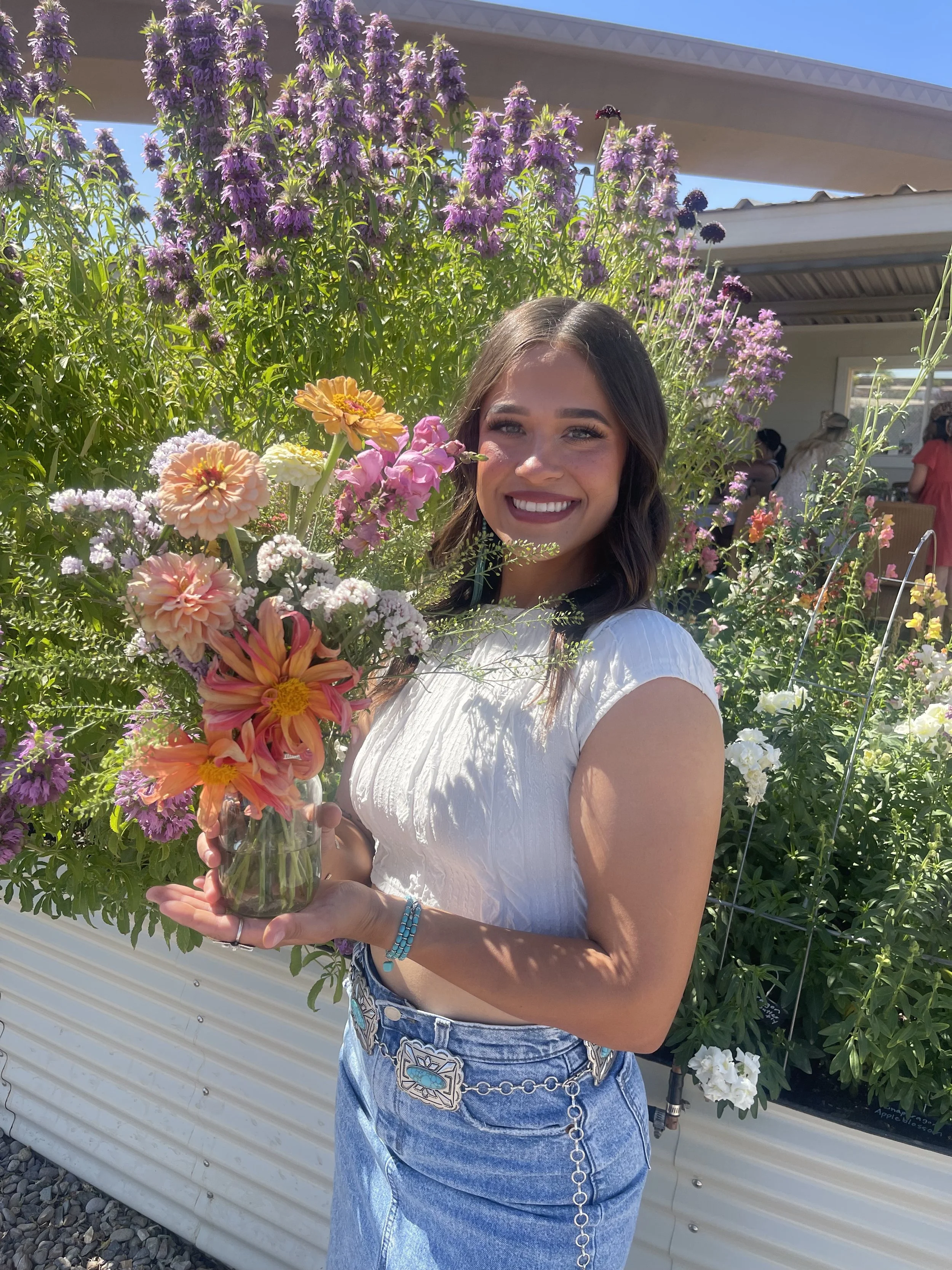 Smiling woman holding a colorful bouquet of flowers in front of a garden filled with purple and white flowers under a clear blue sky.