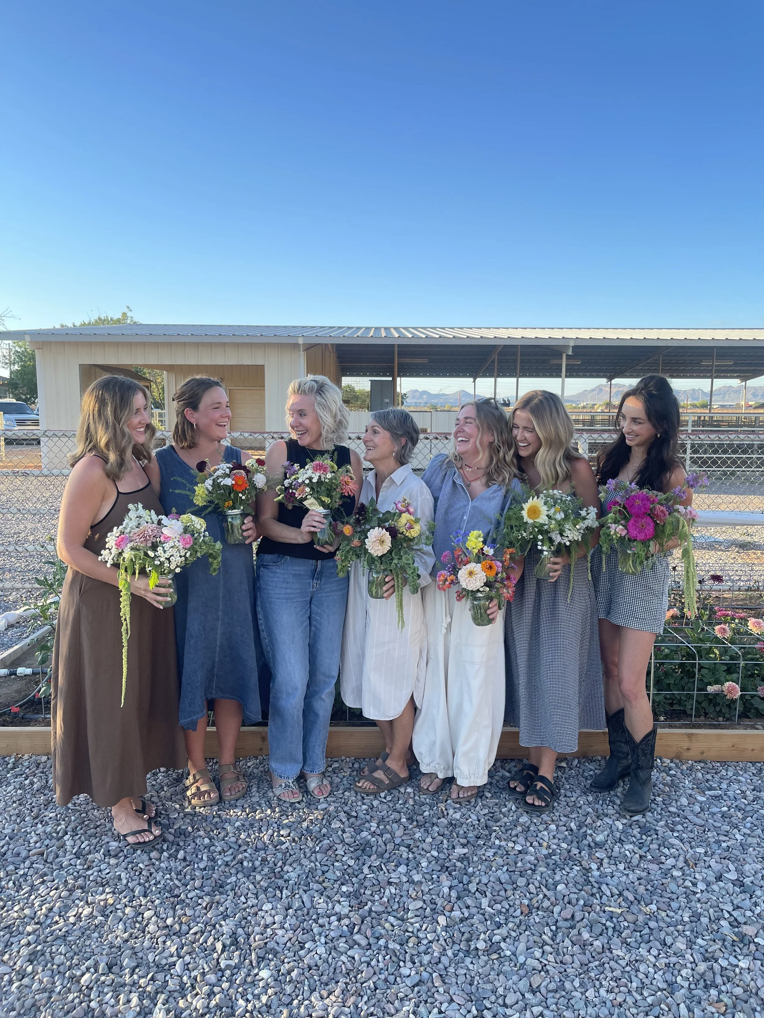 Group of seven women standing outside on gravel with a fence and a small building behind them, holding colorful flower bouquets, smiling and laughing together.