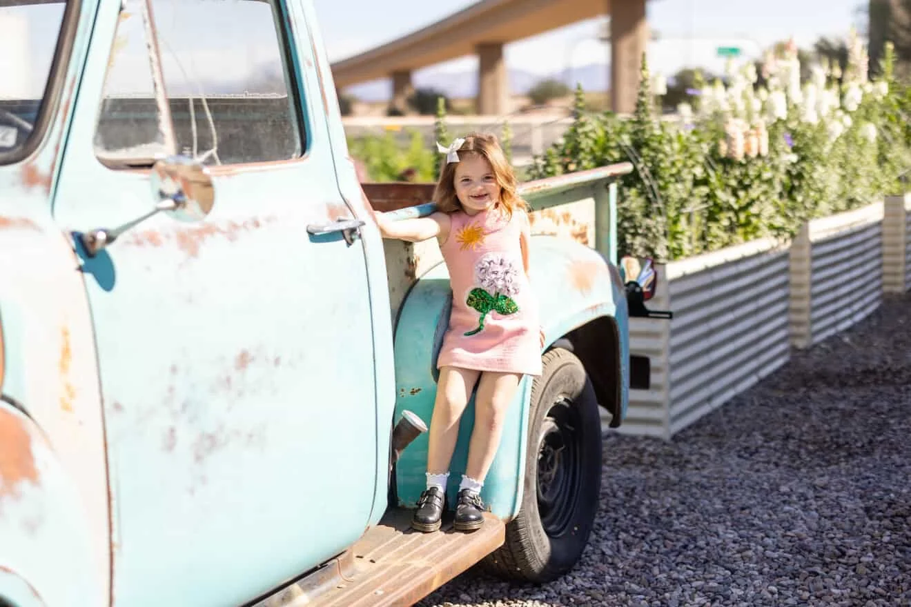 A smiling young girl in a pink dress with a sun, lion, and snake design, standing on the running board of a vintage light blue pickup truck with rust spots. She is outdoors near a flower garden with a fence and an overpass in the background.