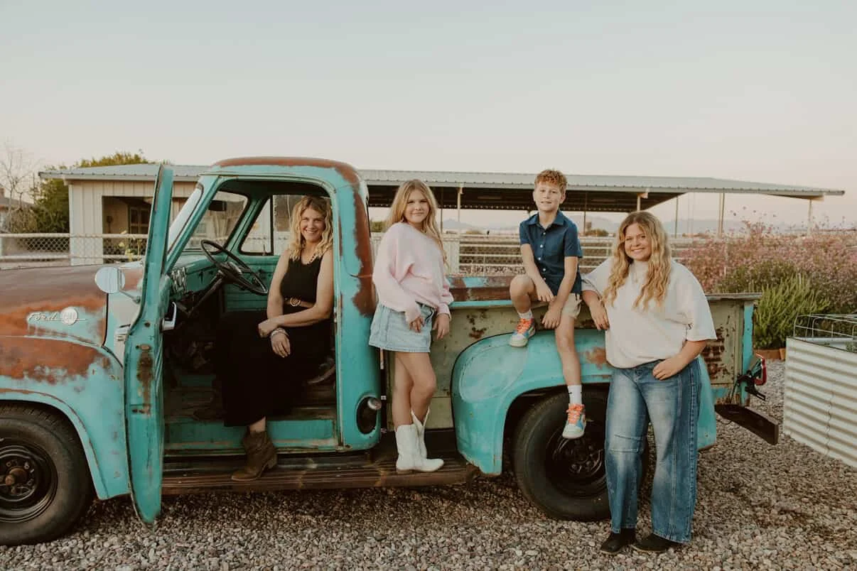 Four people, two women and two children, posing with an old, weathered turquoise pickup truck outdoors during daytime.