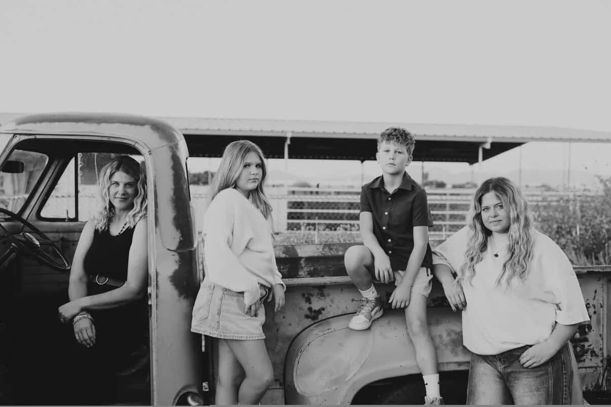 Four young people in casual clothing near an old pickup truck in an outdoor area.