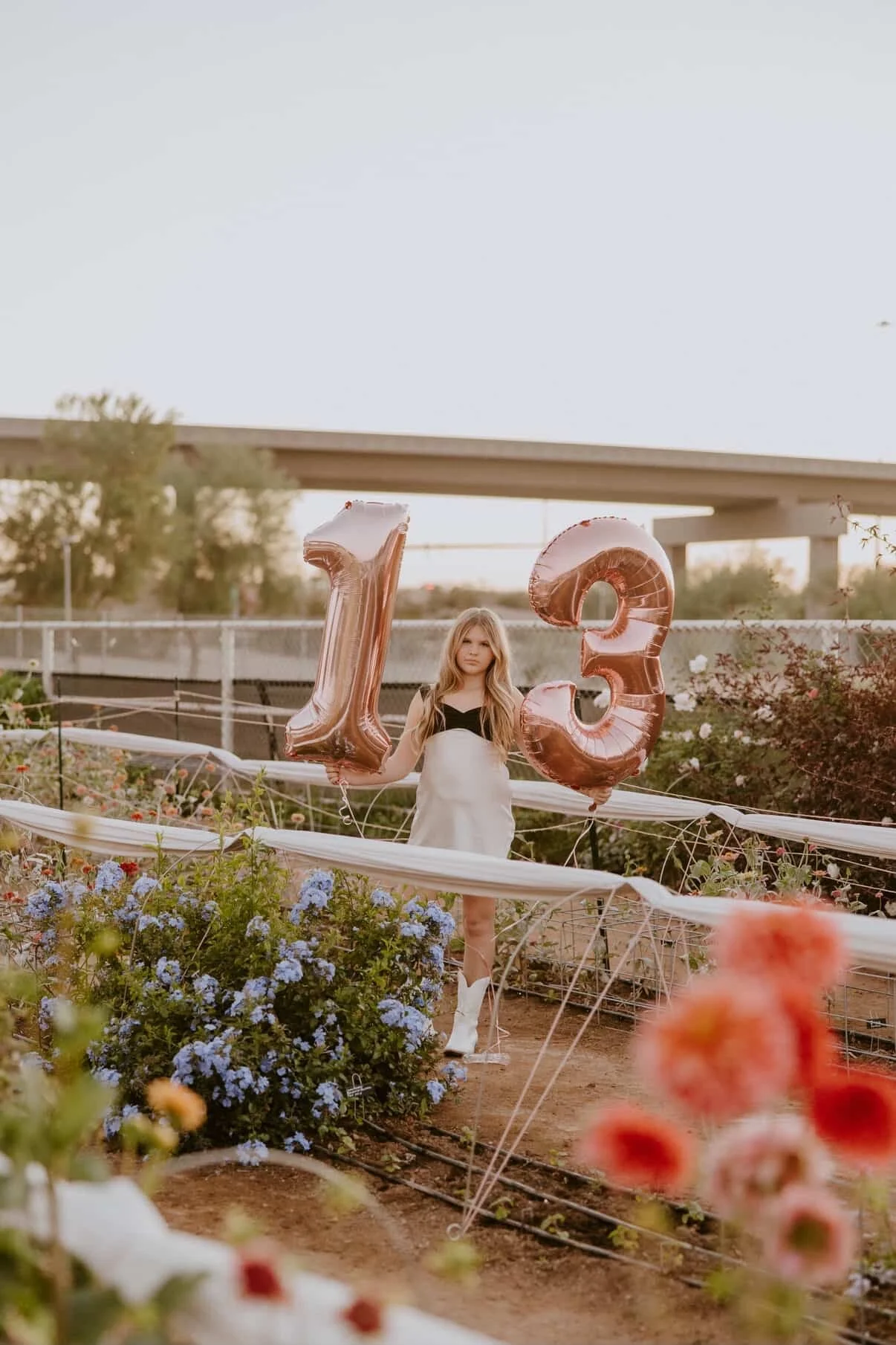 Girl standing in a flower garden holding large rose gold balloons shaped as the numbers 1 and 3.
