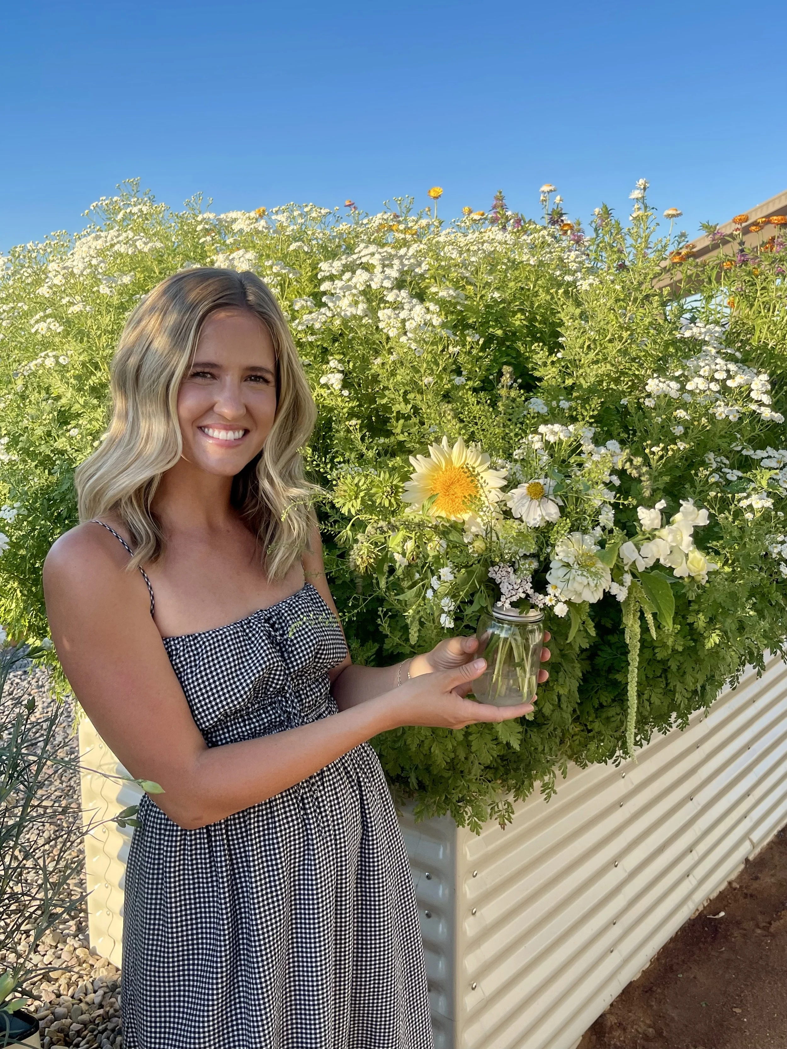 A young woman with blonde wavy hair smiling and holding a small jar with a yellow flower inside, standing next to a large container of white and yellow flowers against a blue sky.