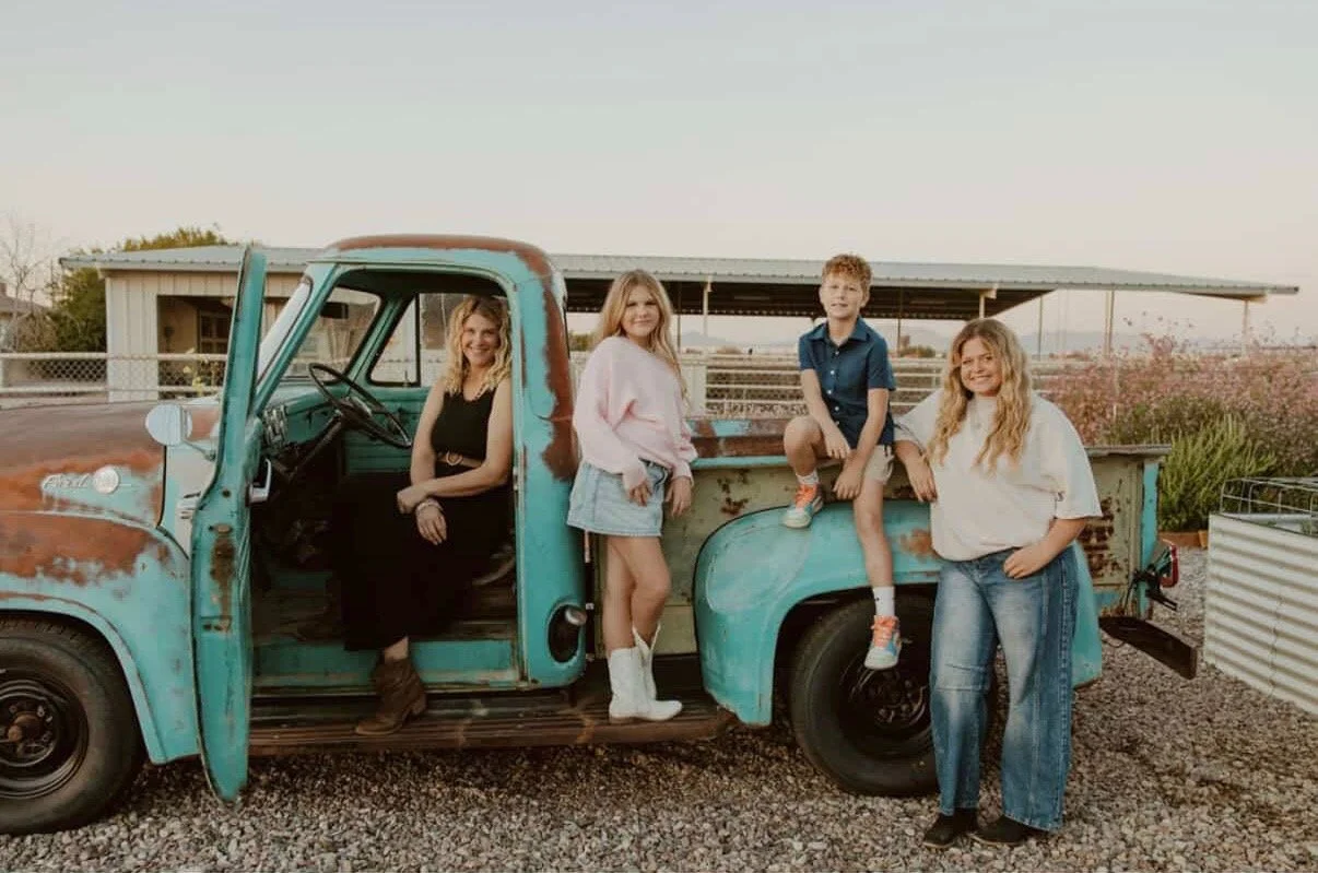 Four people posing with an old, rusted teal pickup truck. The group includes a woman sitting inside the truck, and three children standing and sitting on the truck bed outdoors.