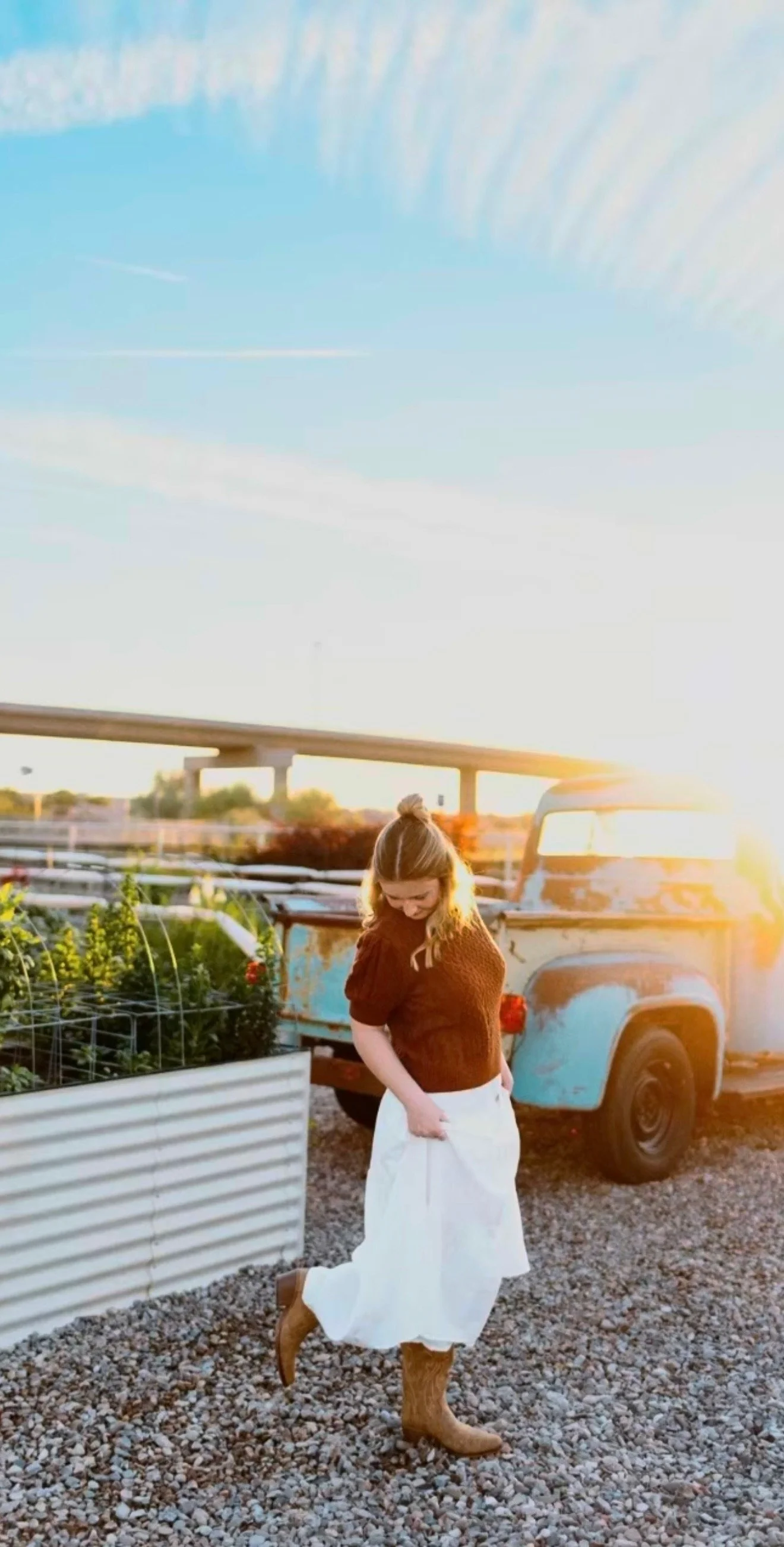 A woman standing outdoors near a vintage truck and a raised garden bed during sunset, wearing a brown sweater, white skirt, and cowboy boots, smiling and looking down.