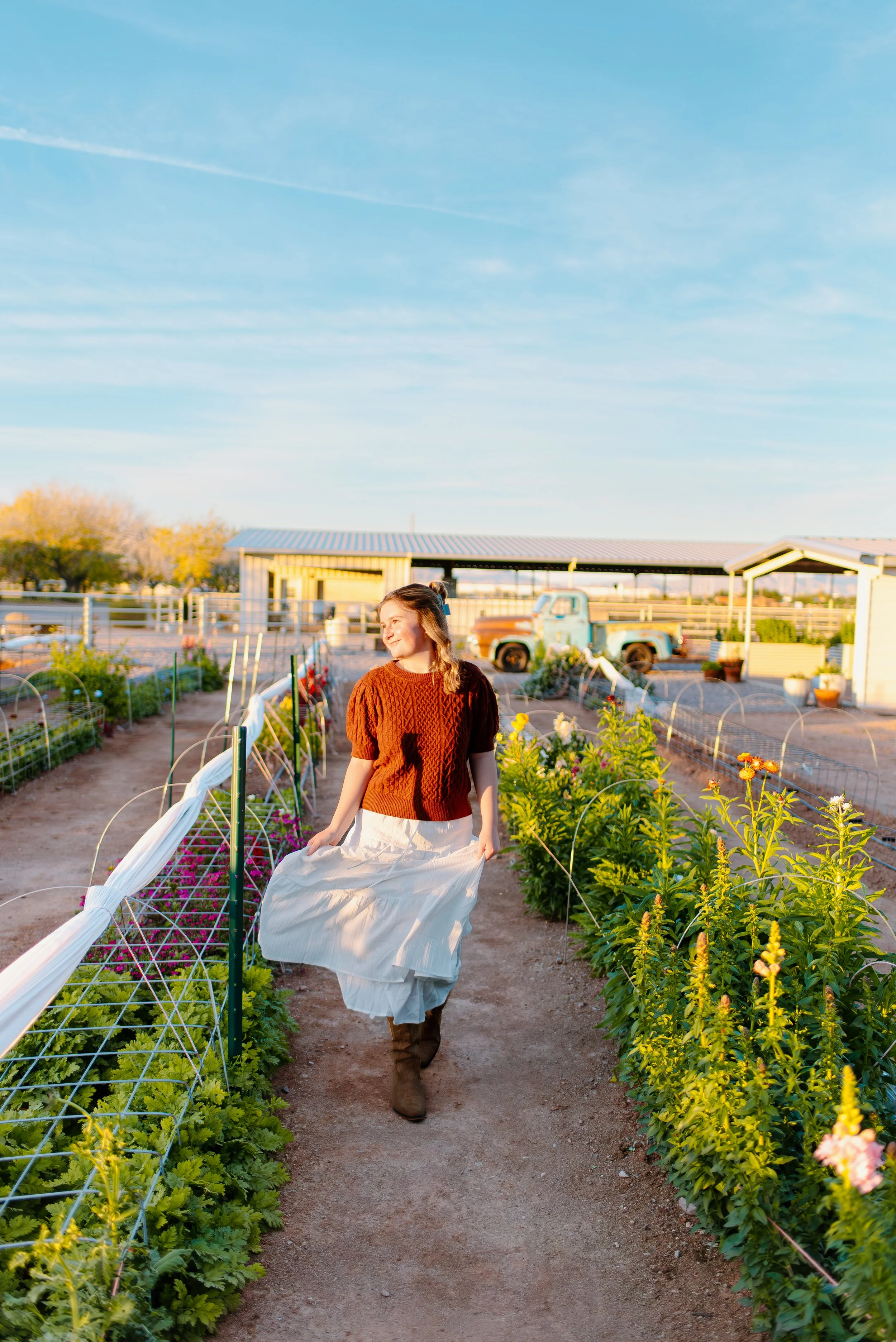 A woman walking through a vegetable garden at sunset, wearing a brown sweater, white skirt, and boots.