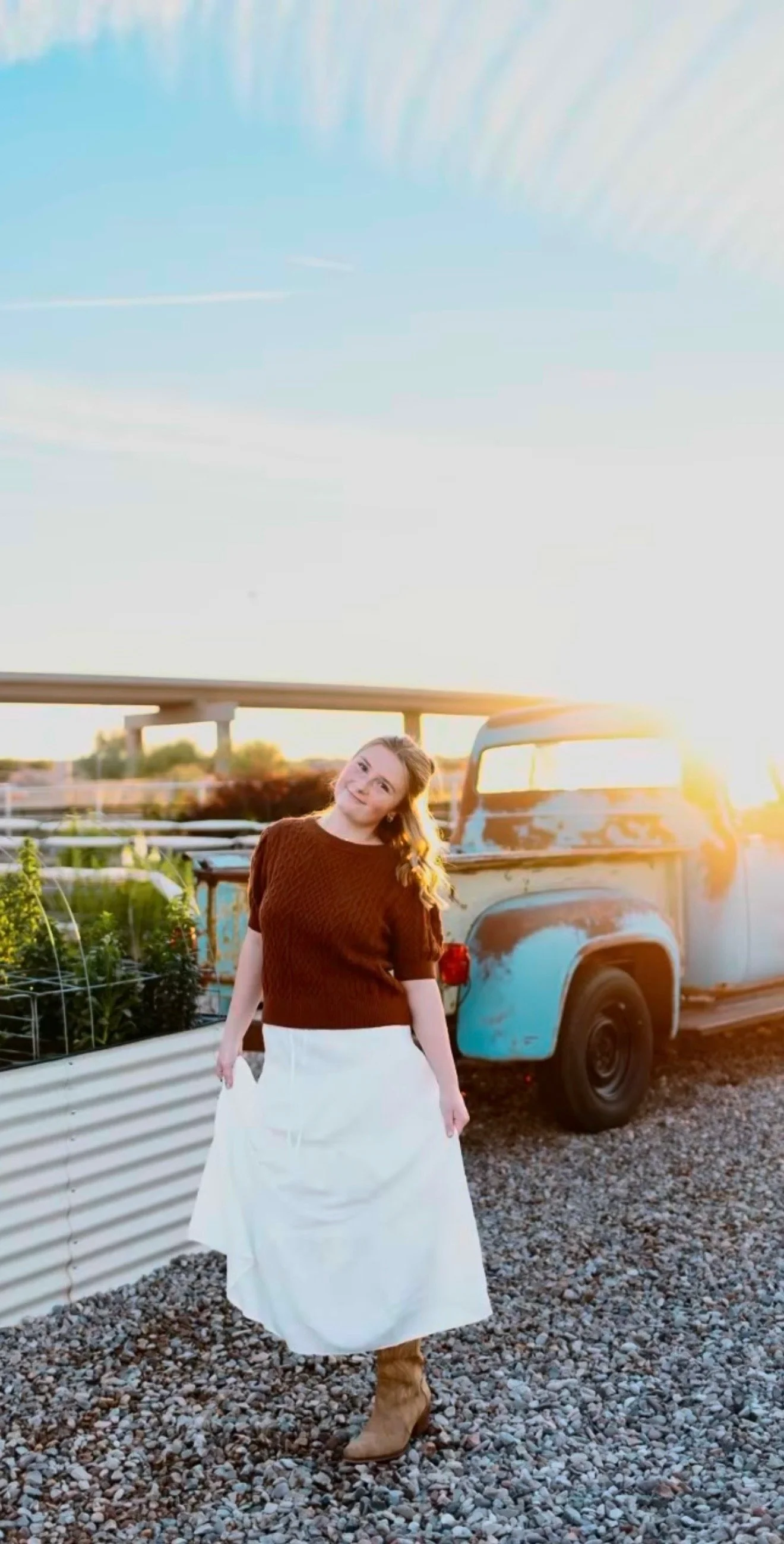 A woman standing on gravel in front of a vintage truck with rust and peeling paint, during sunset. She is wearing a brown sweater, a white skirt, and brown boots, with her hair loose and smiling at the camera.