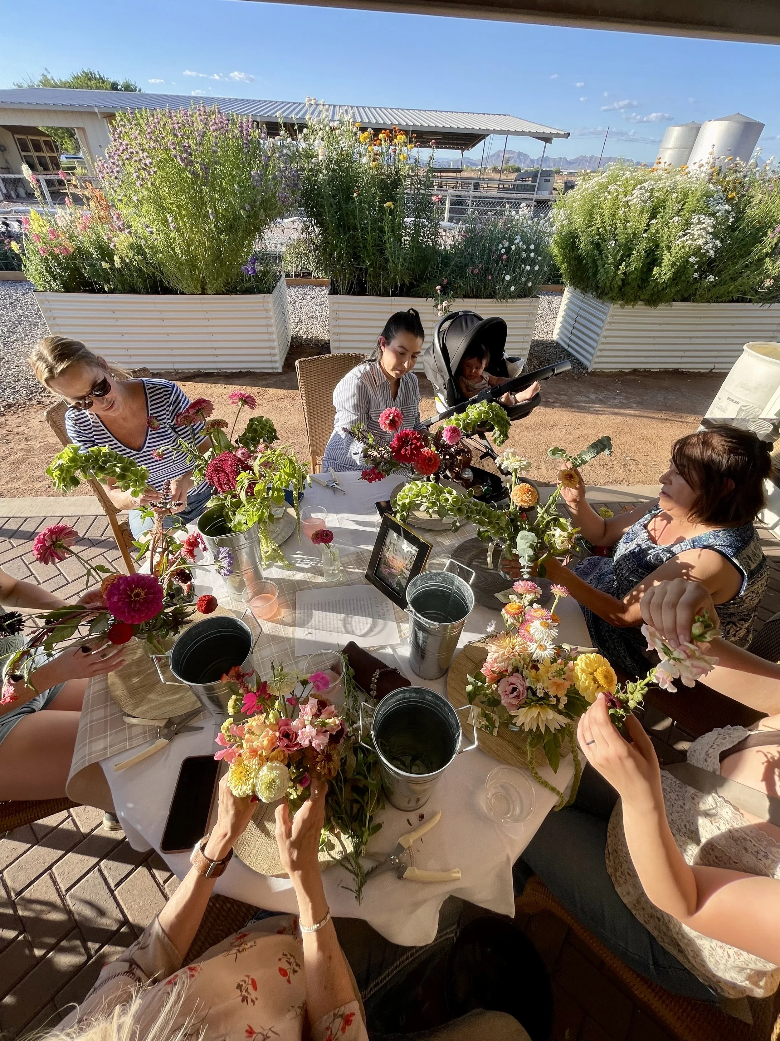 Group of women and children gathered around a table outdoors, arranging colorful flower bouquets with large planters of blooming flowers in the background under a sunny sky.
