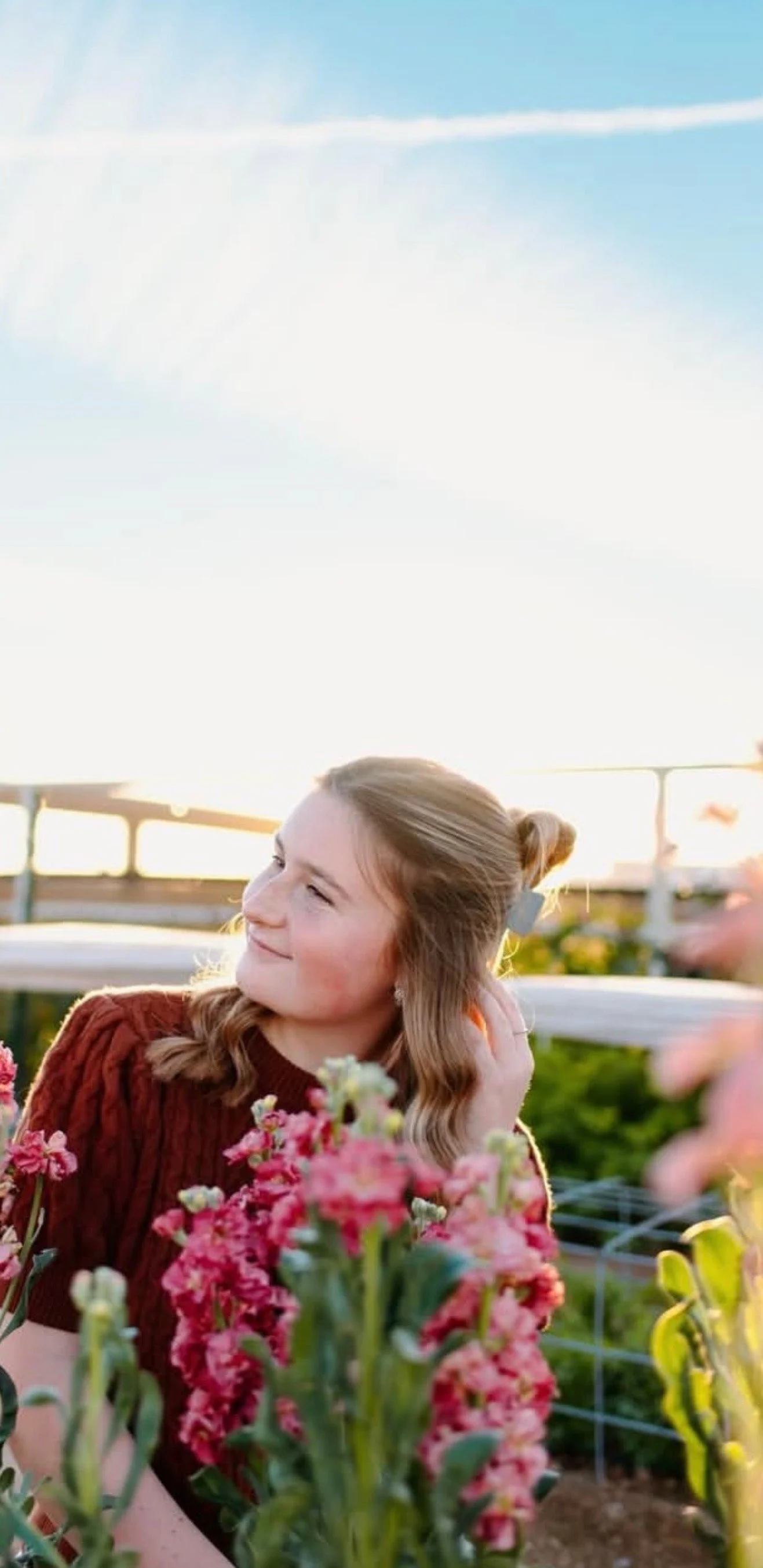 A young woman with brown wavy hair in a bun, wearing a red sweater, standing in a garden with pink flowers during sunset, smiling and looking to the side.