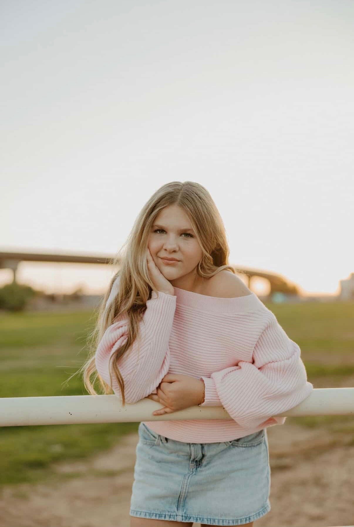 Young woman with long blonde hair wearing a pink sweater and denim skirt, leaning on a white fence outdoors at sunset.