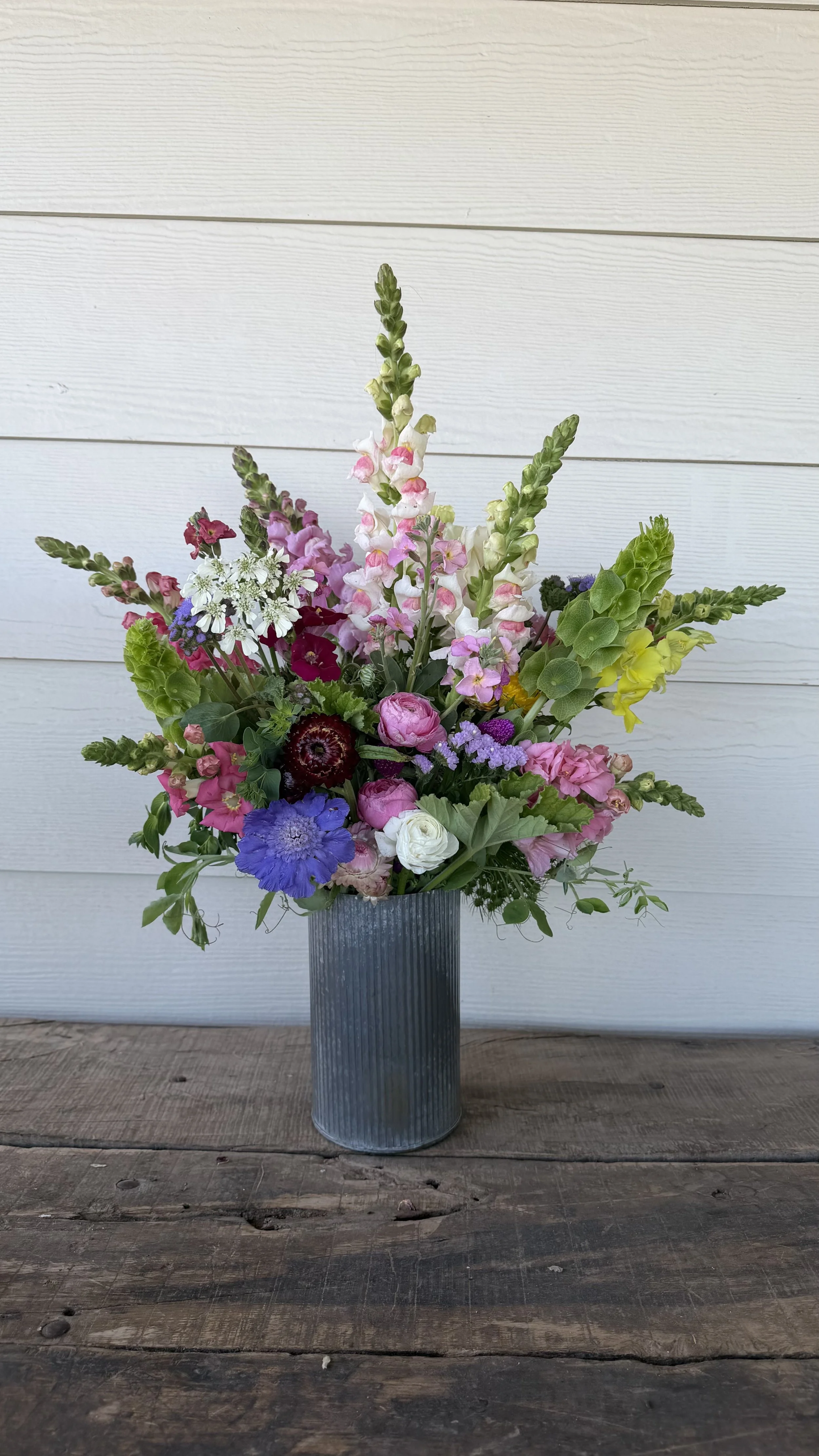 A colorful bouquet of mixed flowers in a gray, ribbed metal vase on a rustic wooden table with a white wall background.