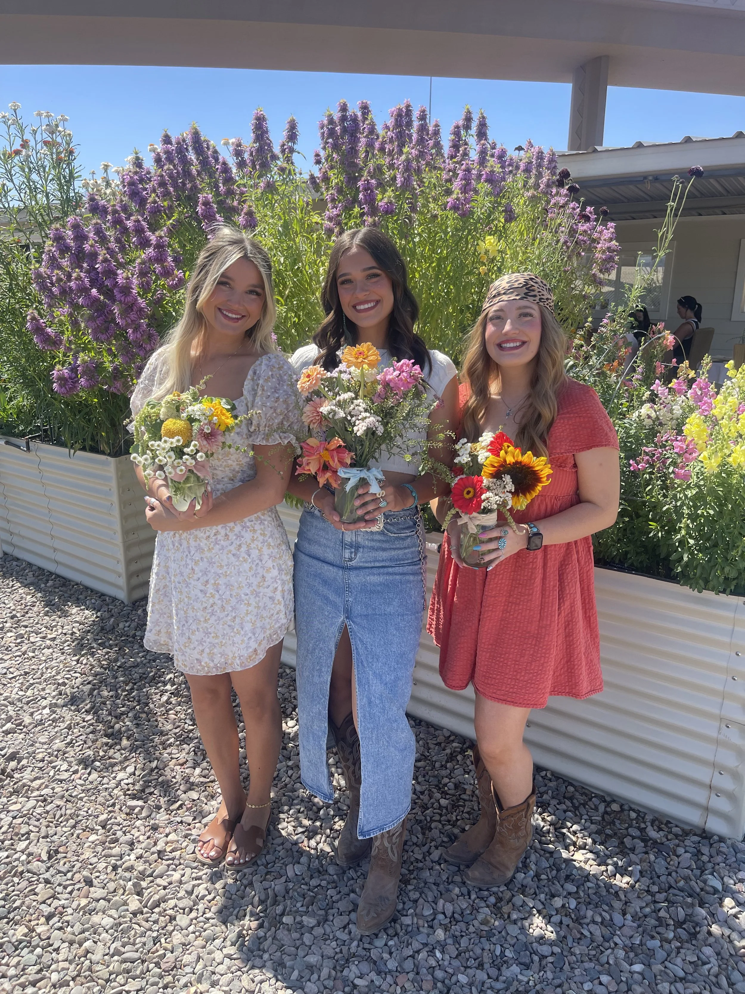 Three women standing outdoors in front of a large flower bed, holding bouquets of flowers, smiling at the camera on a sunny day.