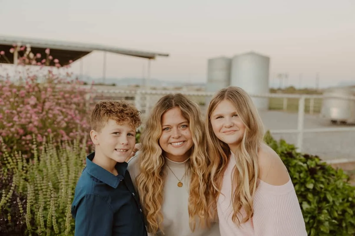 A woman with long curly hair and a white shirt smiling alongside a boy with short curly hair in a blue shirt and a girl with long wavy hair in a light pink off-shoulder top, standing outdoors with pink flowers and buildings in the background.