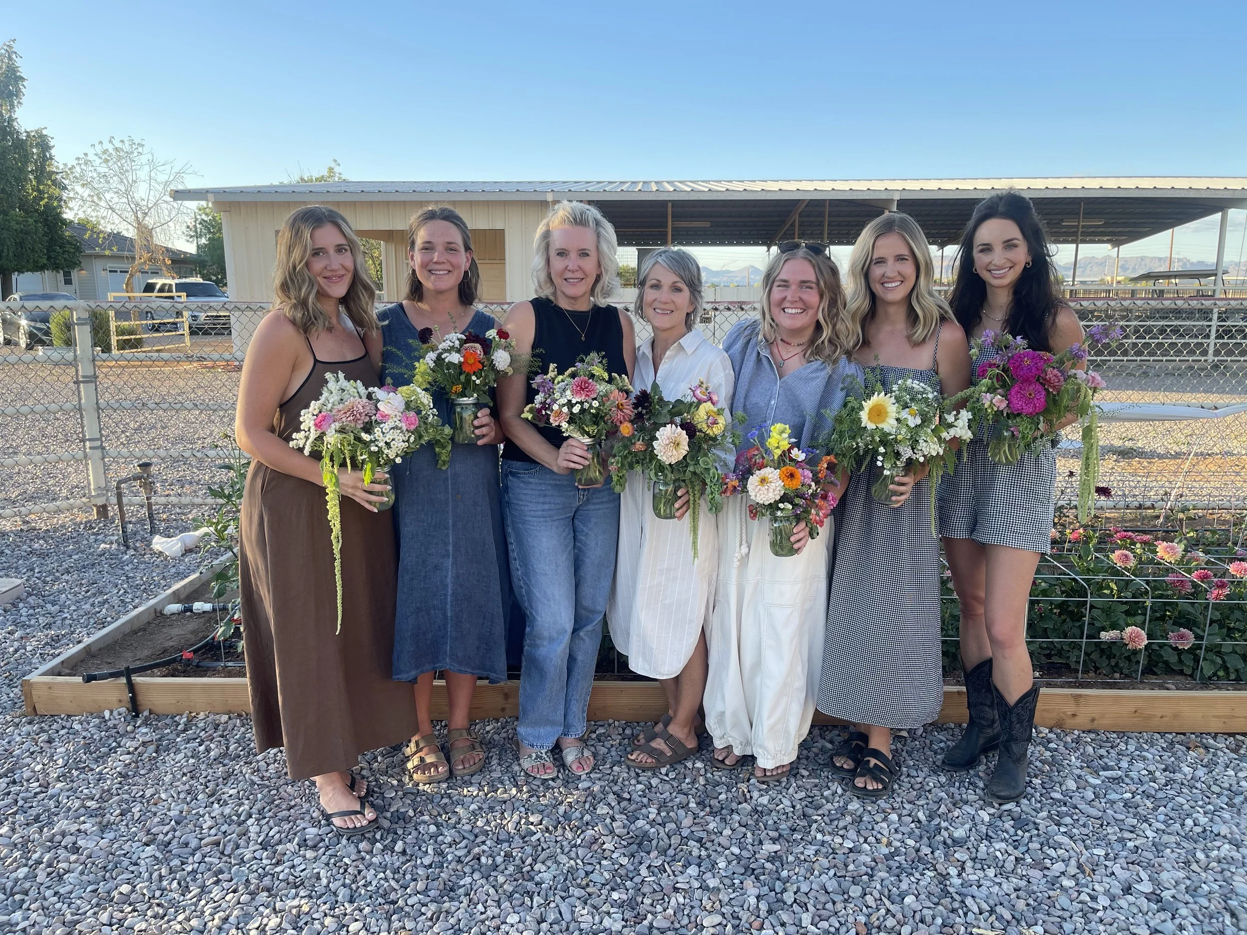Seven women standing outdoors in front of a fence, each holding a bouquet of colorful flowers, smiling for the camera on a sunny day.