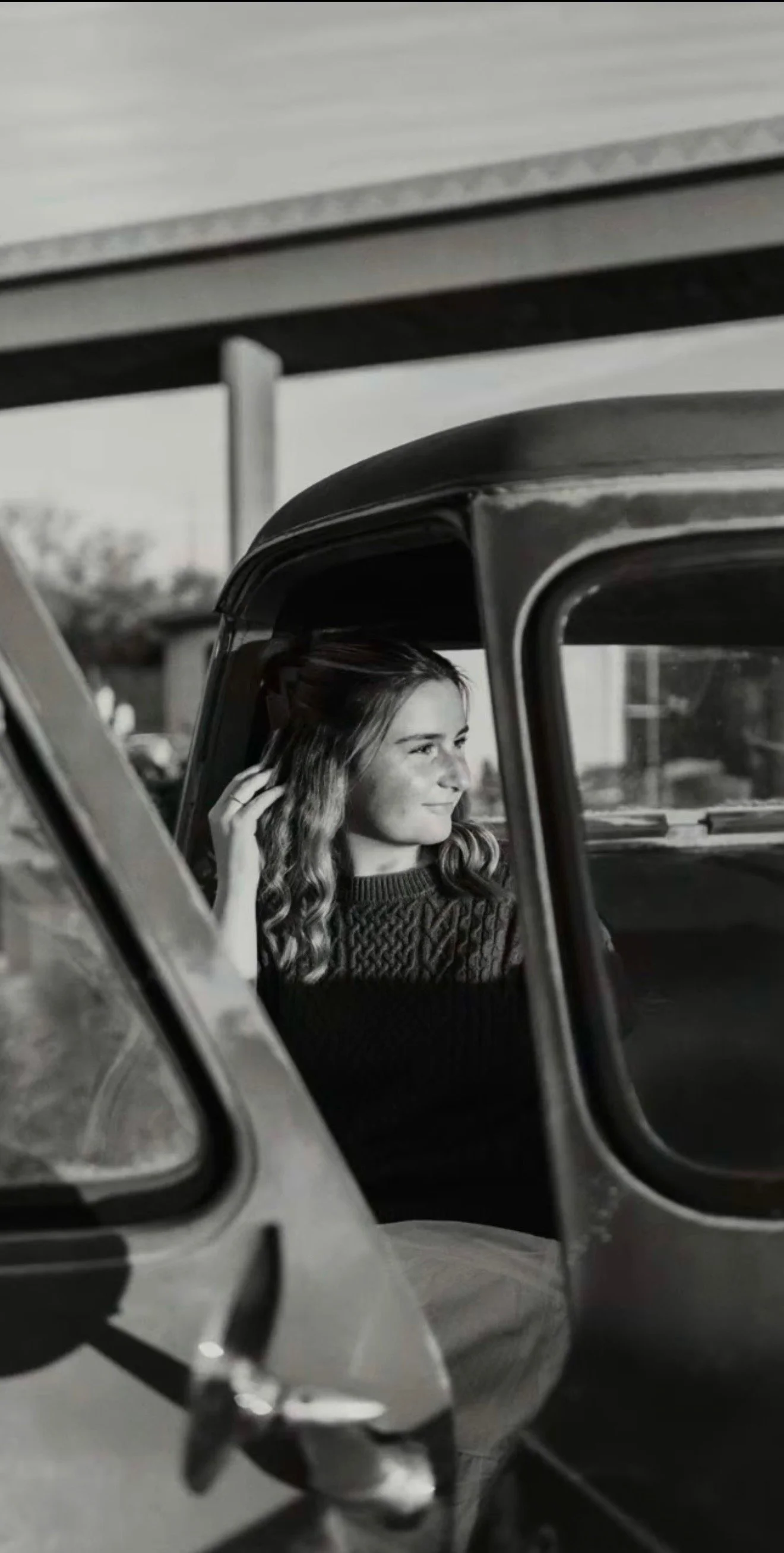A young woman with wavy hair sitting inside a vintage car, smiling and looking out the window.