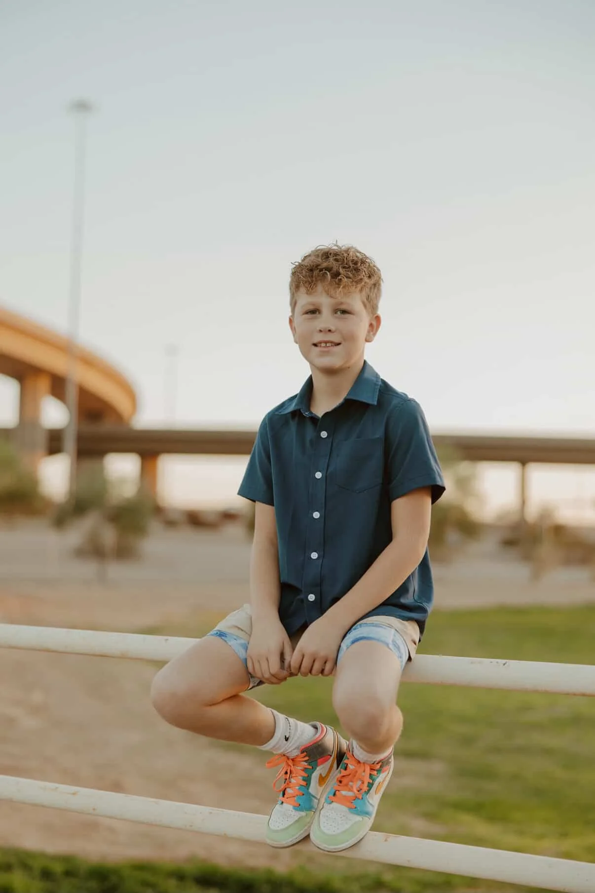A young boy with curly red hair, wearing a blue short-sleeved button-up shirt, beige shorts, and colorful sneakers, is sitting on a white fence near a park or outdoor space with a bridge in the background.