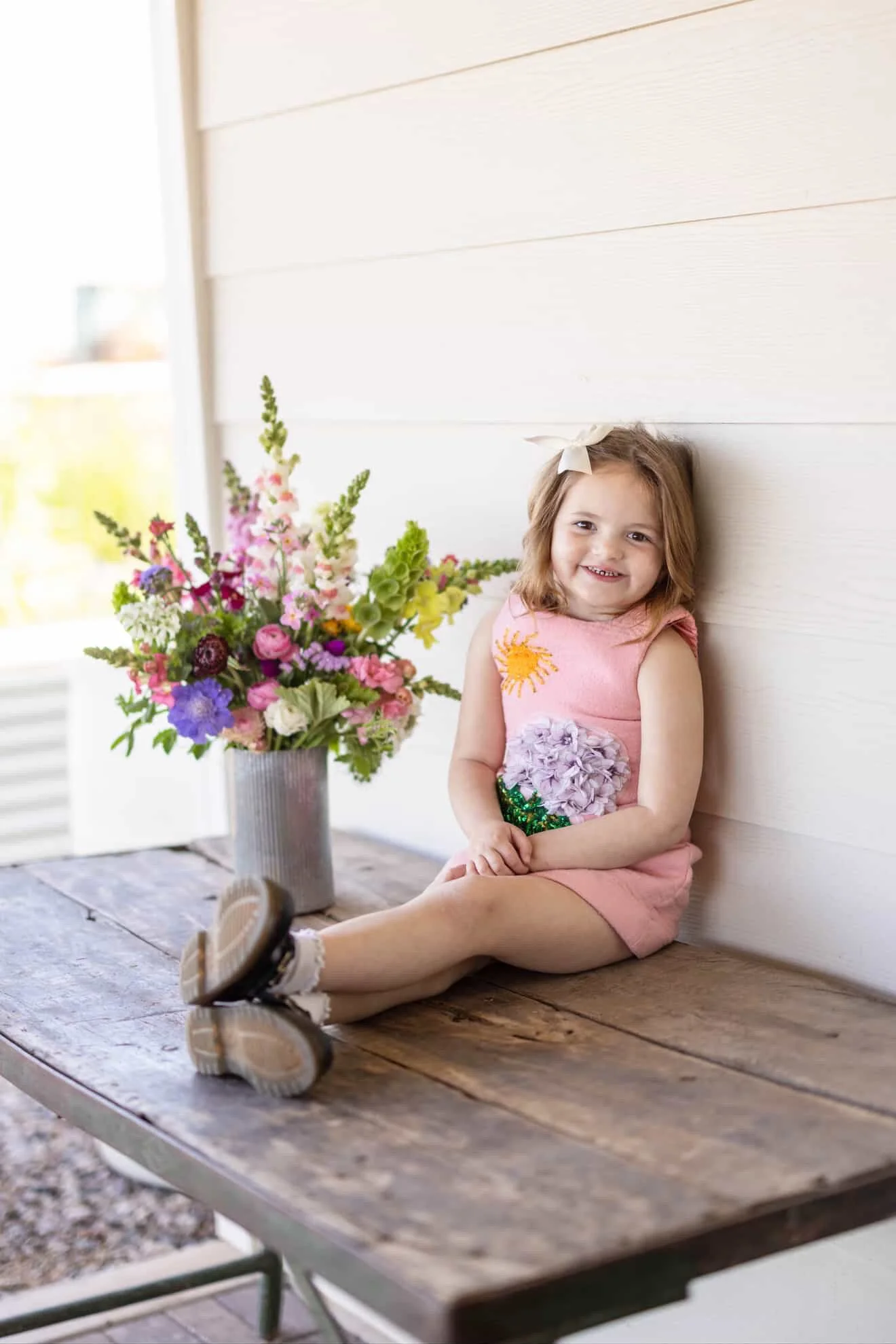A young girl sitting on a wooden table, smiling, with a large colorful bouquet of flowers in a gray vase beside her, against a white wall.