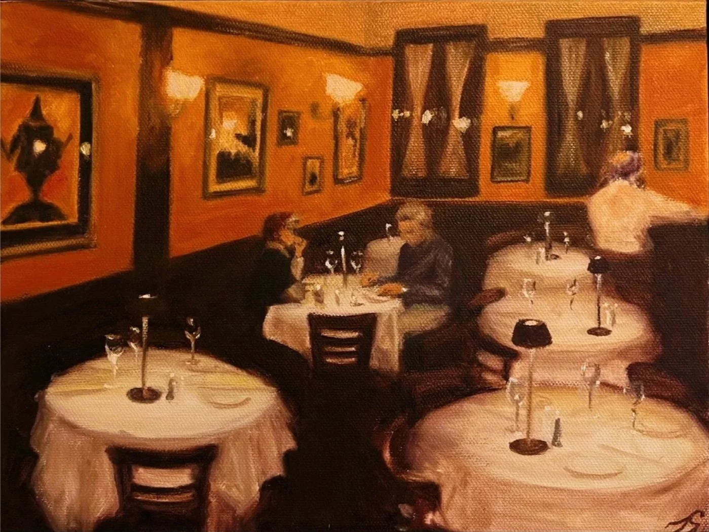 A restaurant interior with orange walls decorated with framed paintings, several tables with white tablecloths set with glasses and candles, and a waiter attending a table in the background. Two women are seated at one table engaged in conversation.