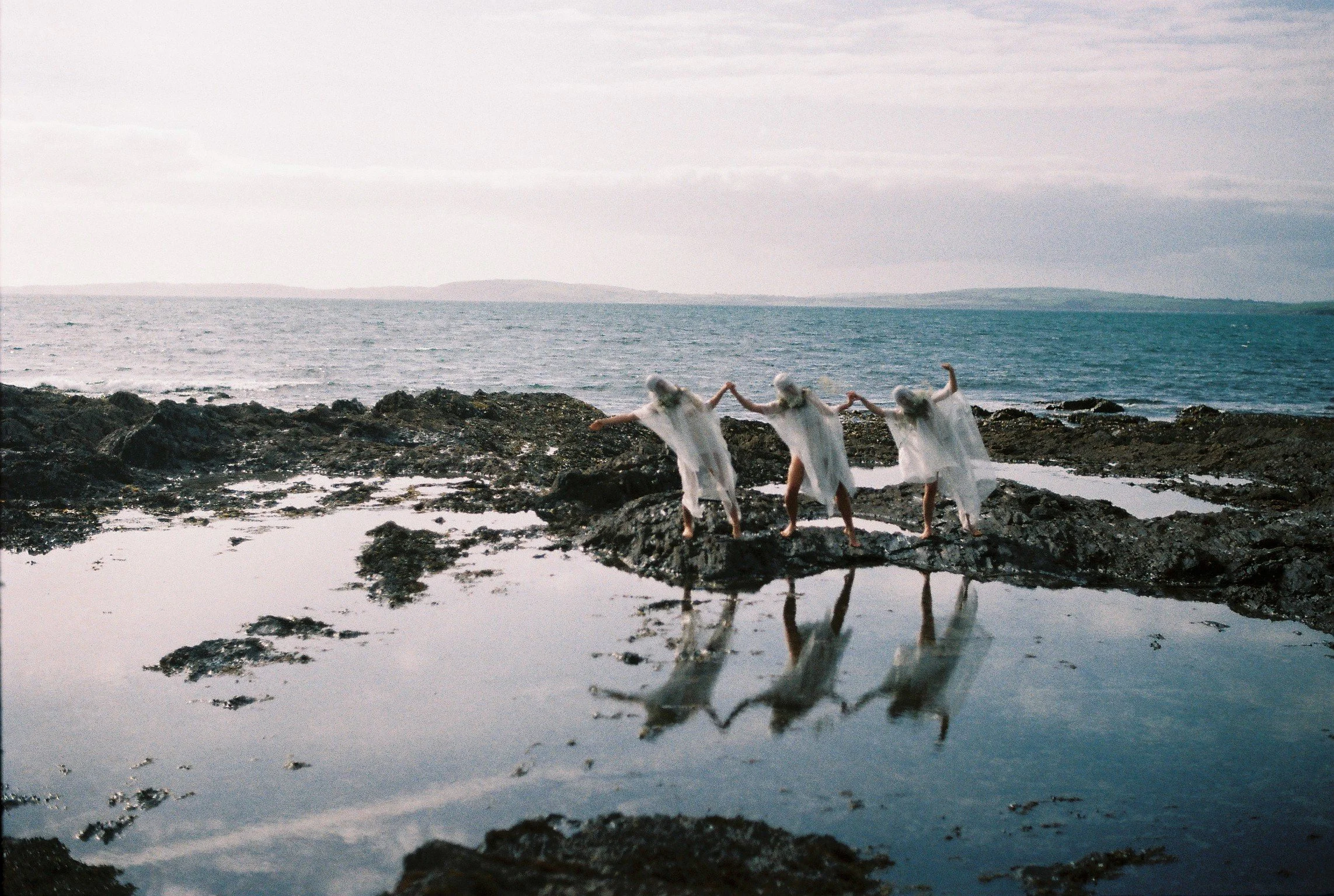 Three figures dressed in flowing white garments dancing on rocky shoreline near the ocean, with their reflections visible in a tidal pool in the foreground.