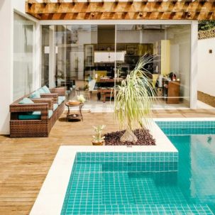 Modern backyard with a swimming pool, bamboo-style wicker sofa with teal cushions, potted plants, and a wooden deck, front of a house with large glass window and sliding door