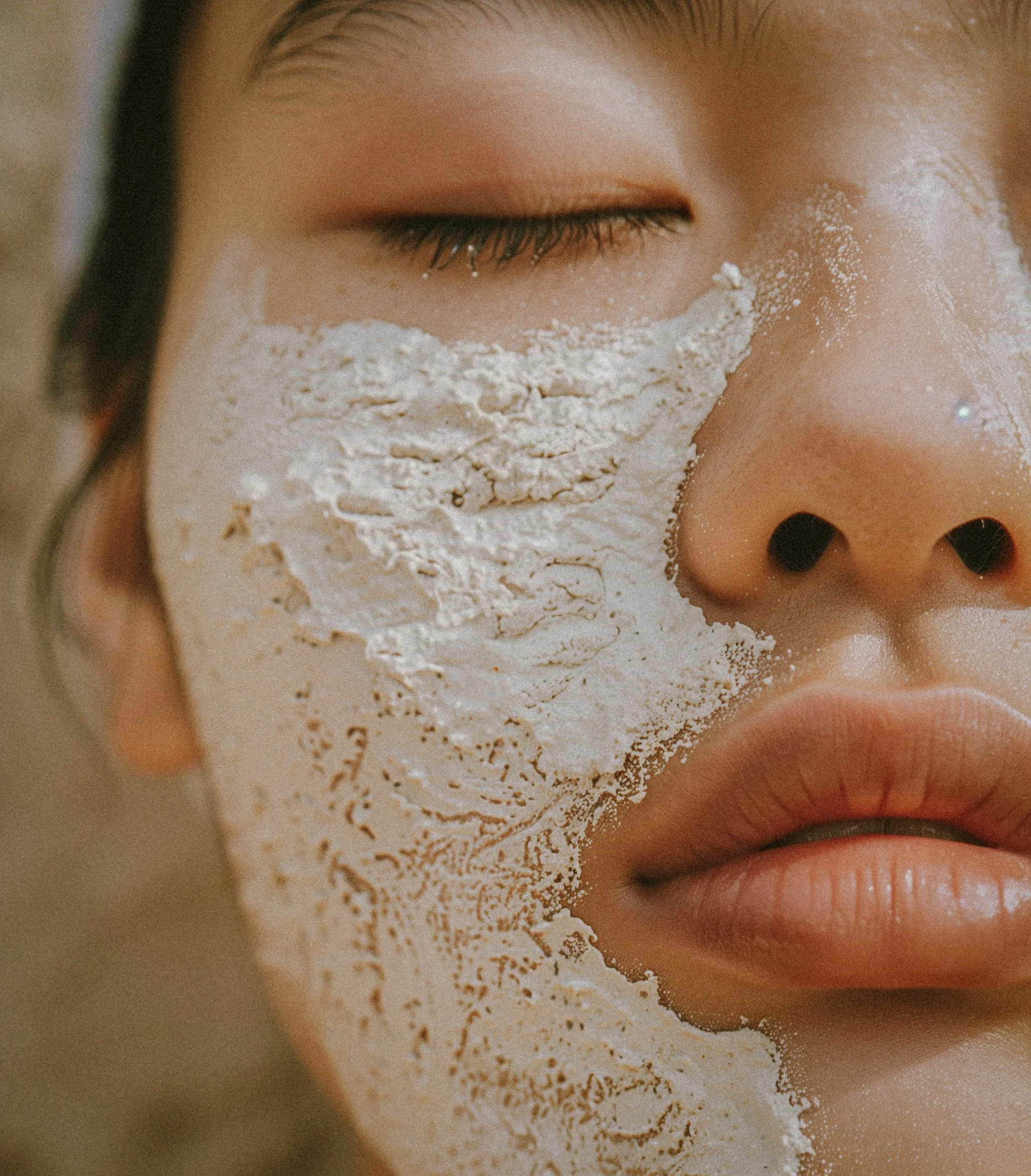 Close-up of a woman's face with cream or mask applied to the right side of her face, covering her cheek and part of her nose, with her eyes closed and lips slightly parted.