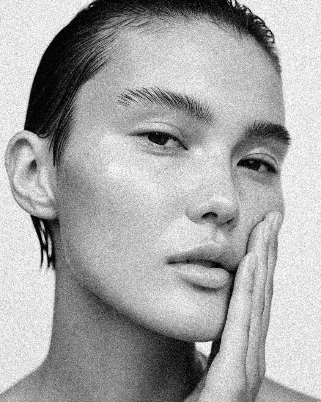 Close-up black and white photo of a woman gently touching her face with her hand, showing smooth skin, arched eyebrows, and short wet hair.