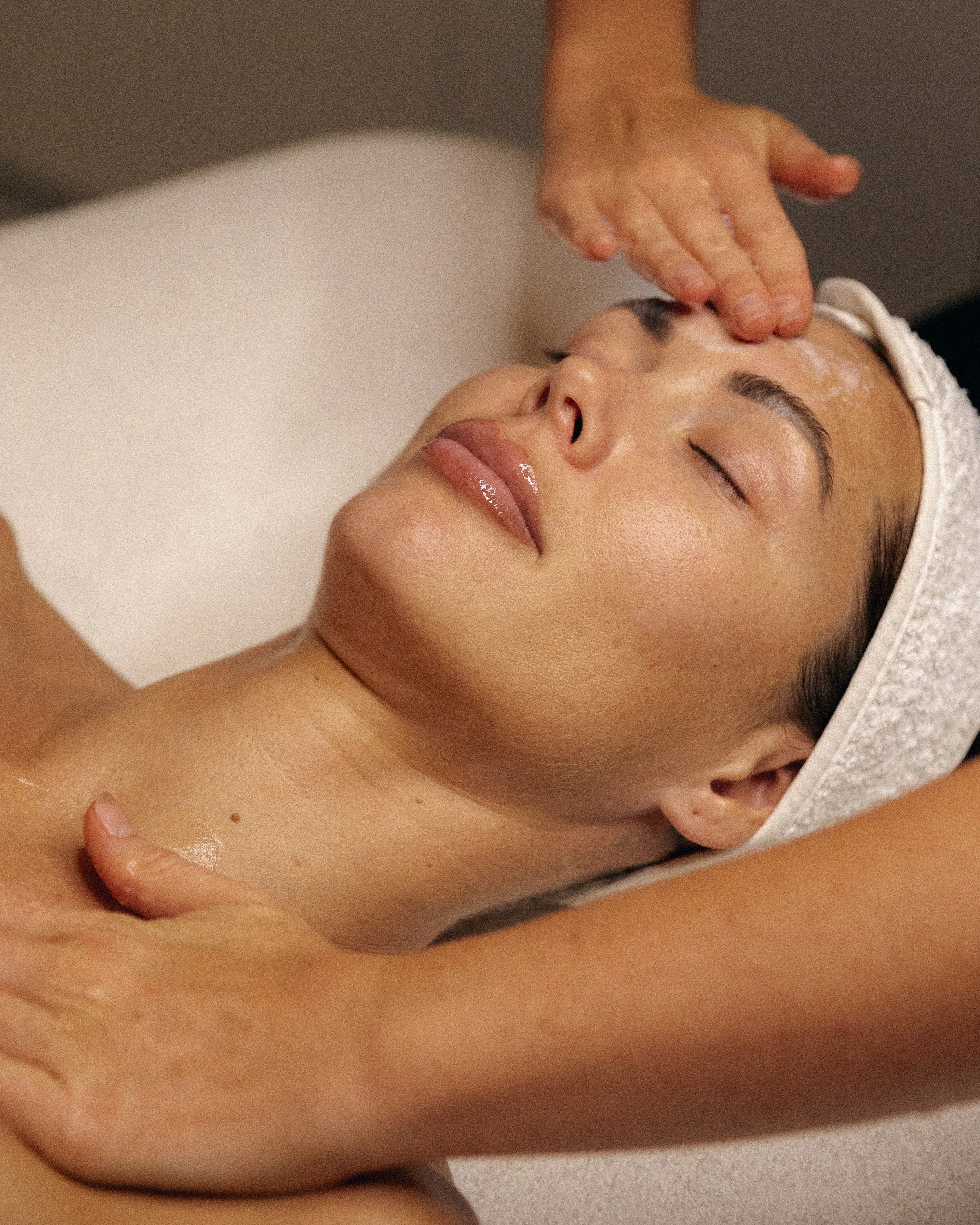 A woman receiving a facial treatment while lying down with eyes closed, a white headband covering her hair, and a person applying a product on her forehead.