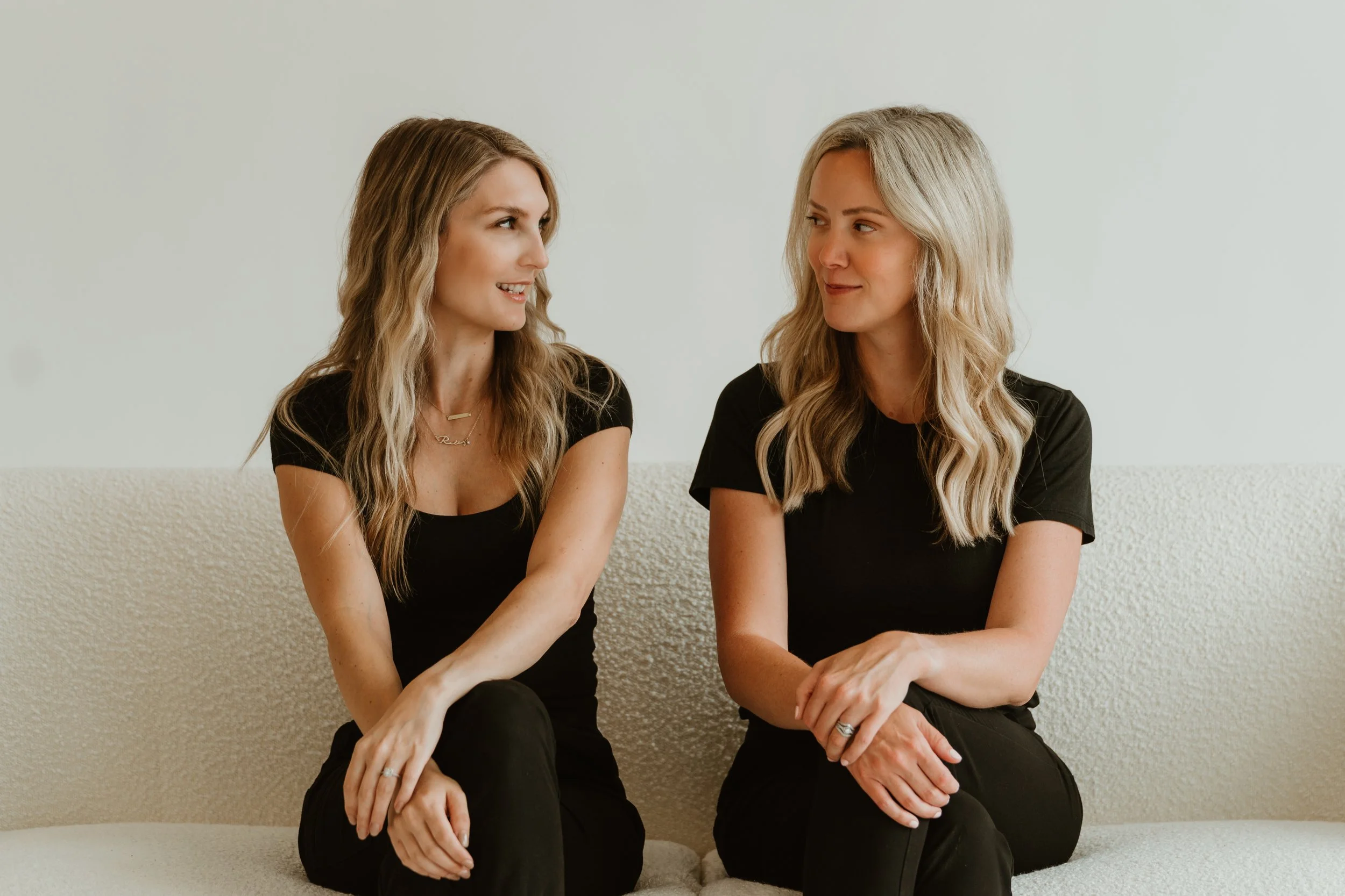 Two women with blonde hair sitting on a beige textured couch, engaging in conversation against a plain off-white wall, both dressed in black tops and pants.