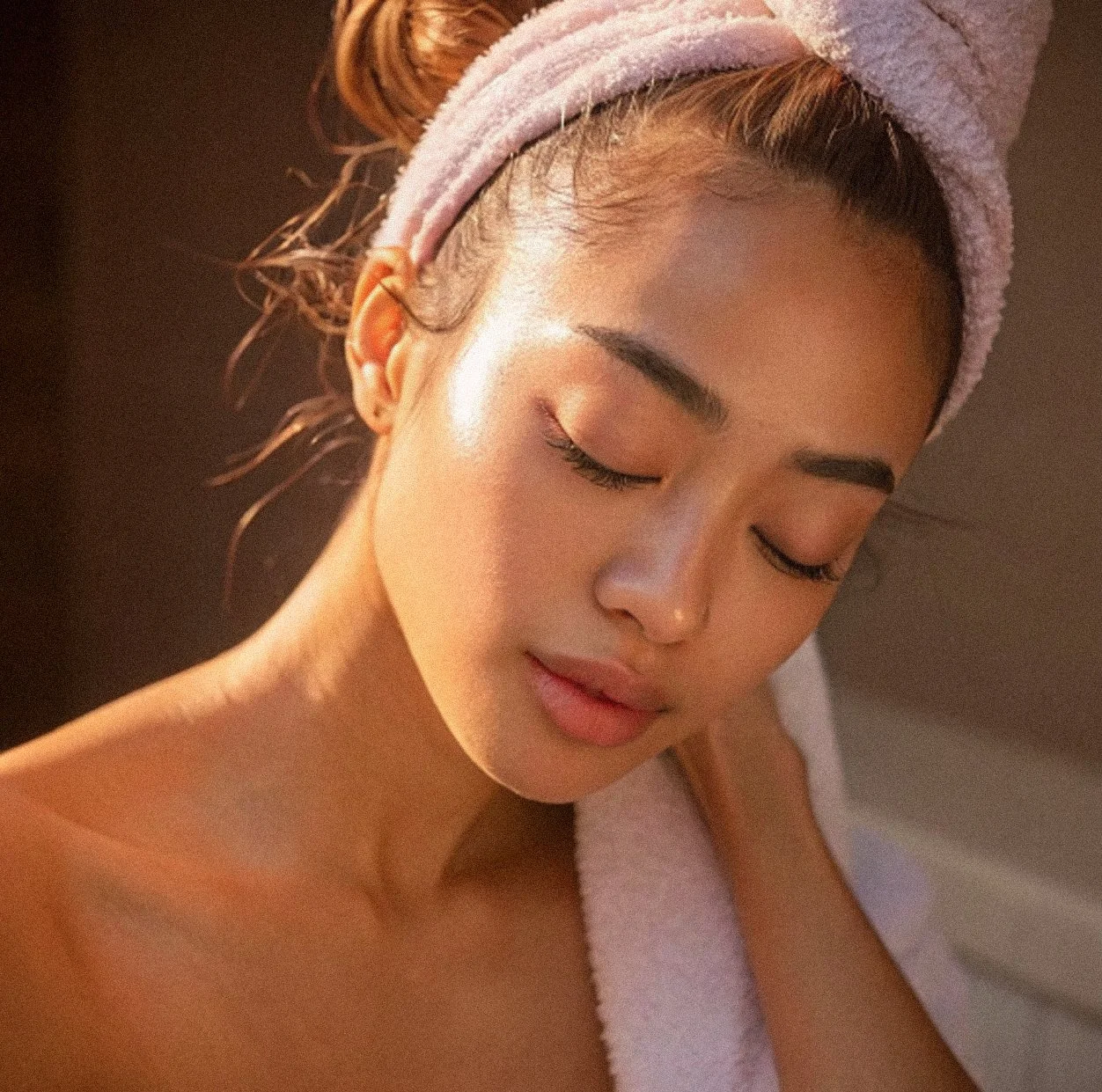 A close-up of a woman with her eyes closed, wrapped in a towel with a pink headband, likely after a spa or skincare treatment.