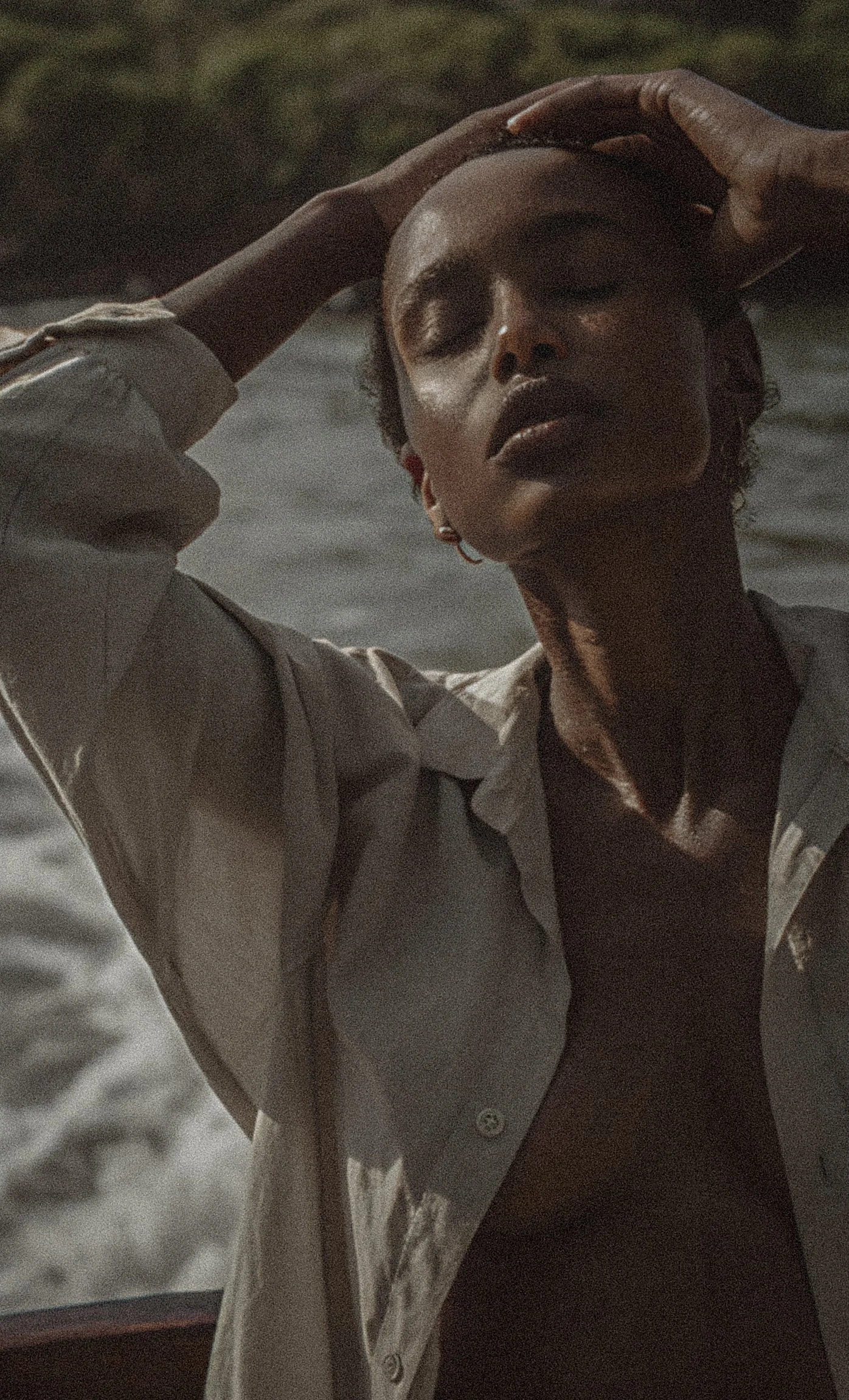 A woman with closed eyes and dark lipstick outdoors, wearing a light-colored shirt with partially unbuttoned collar, touching her forehead with one hand.