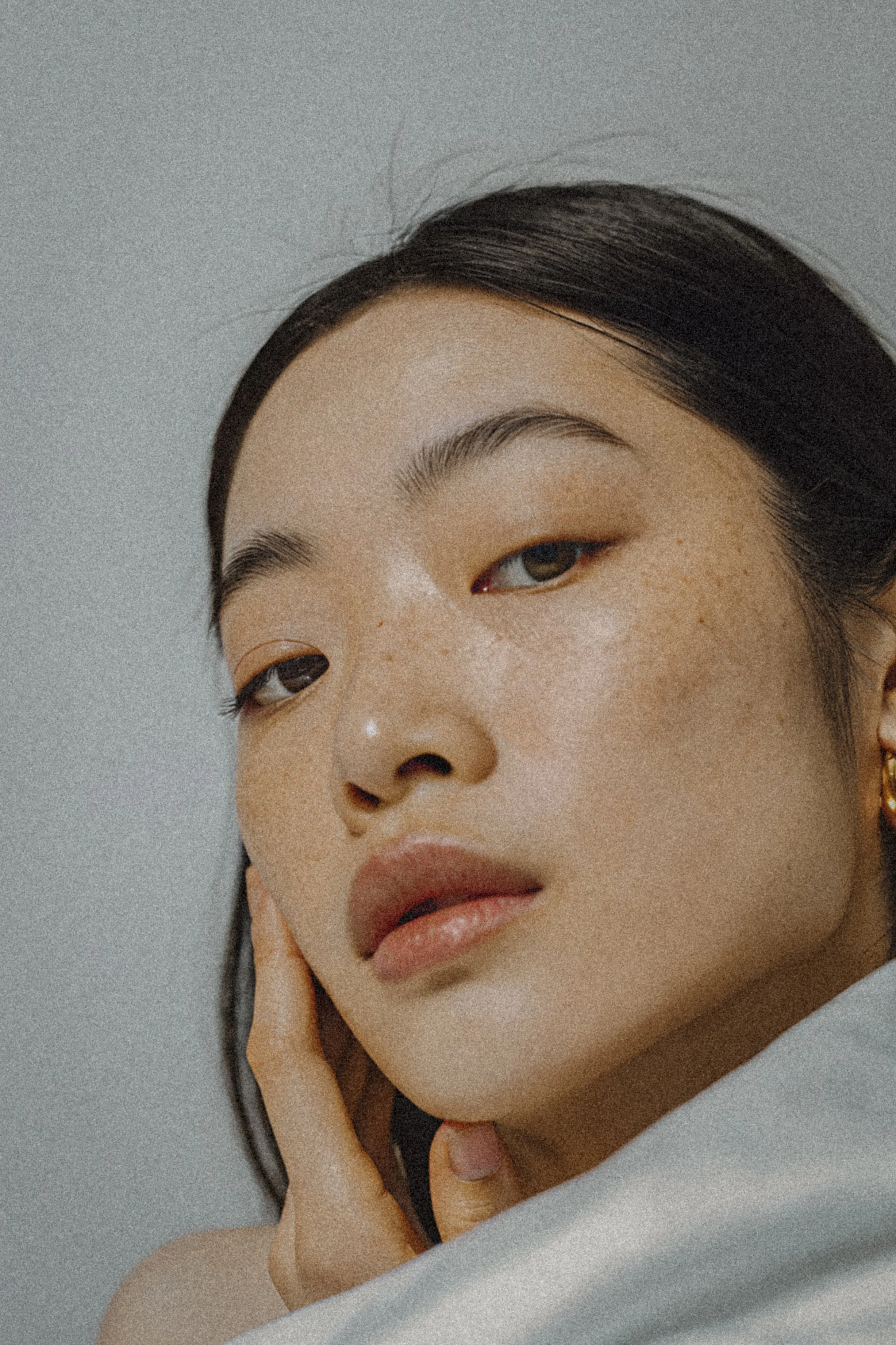 Close-up of a young woman's face with dark hair, freckles, and light makeup, resting her chin on her hand.